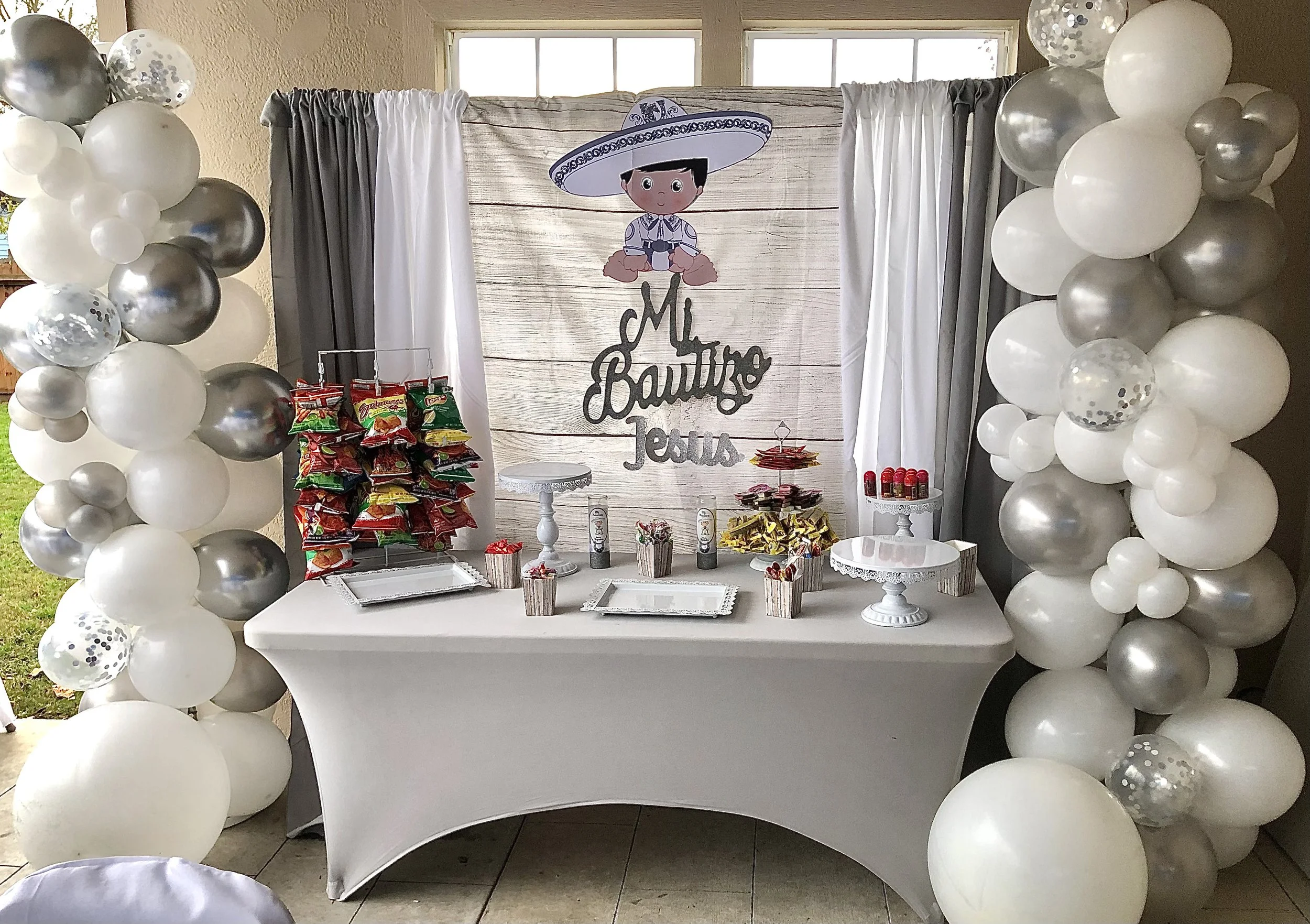 Baby shower decoration featuring a white table with snacks, surrounded by big white, silver, and confetti balloons, a wooden backdrop with a cartoon baby dressed as a farmer complete with a large sombrero, and a sign that reads 'Mi Bautizo Jesus.'