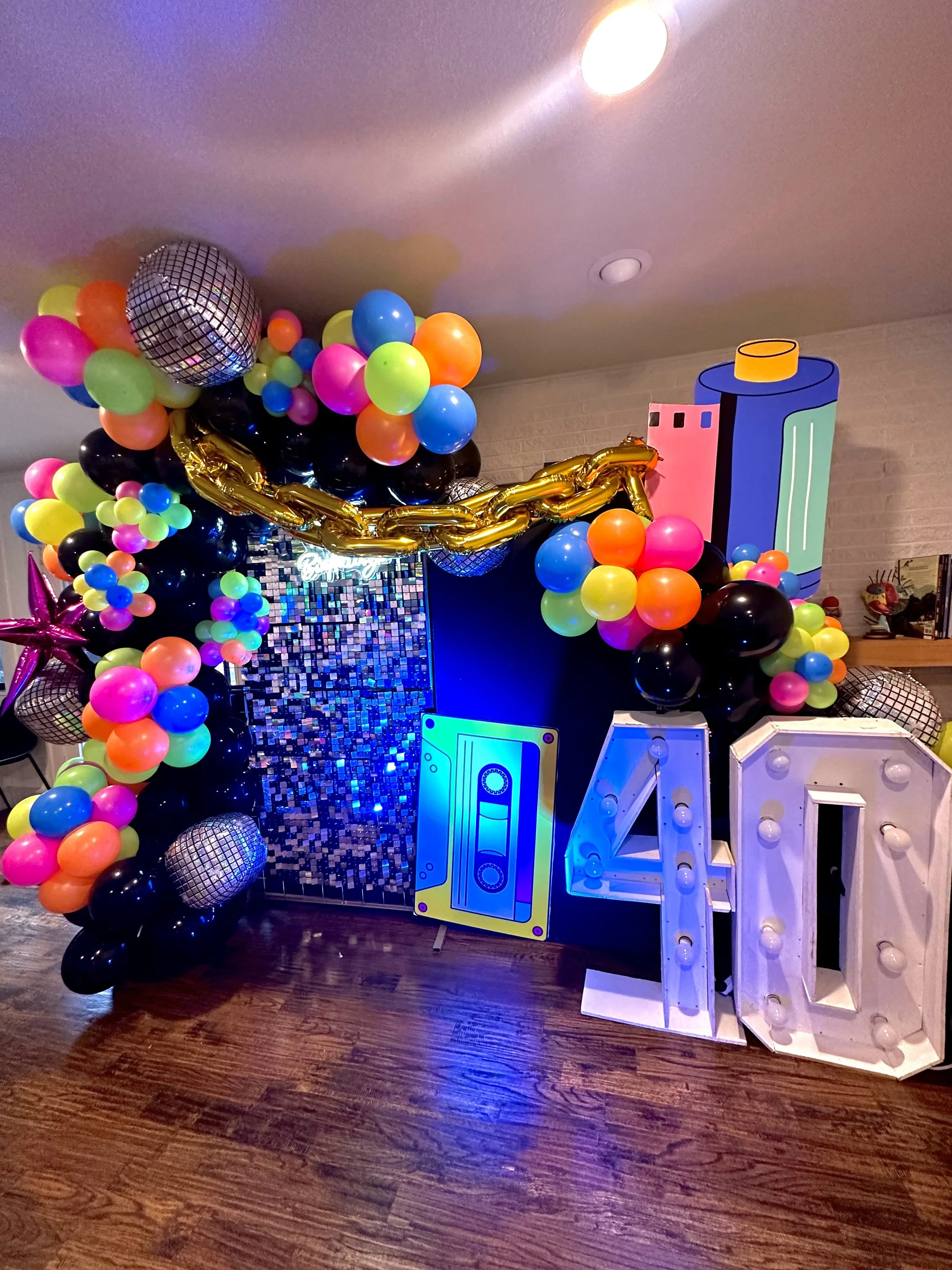 Colorful birthday party decorations including a balloon arch with multicolored balloons, a large disco ball, a twisted gold balloon garland, illuminated letter sign, and a cardboard cutout of a cassette tape. The space has wooden flooring and a brick