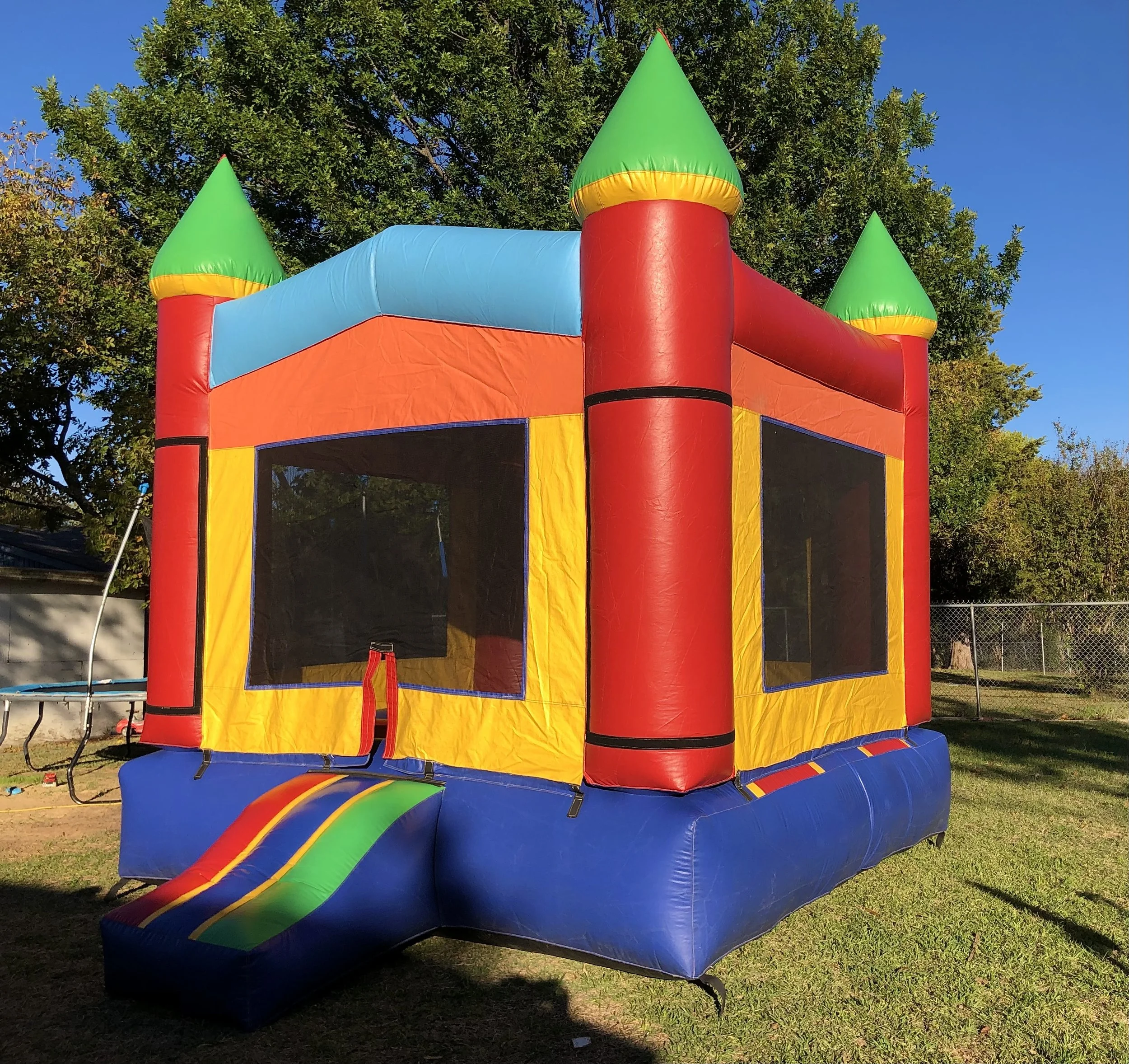Colorful inflatable bounce house with green, red, blue, yellow, and orange sections, featuring cone-shaped turrets and a slide, set outdoors against a background of trees and a clear blue sky.