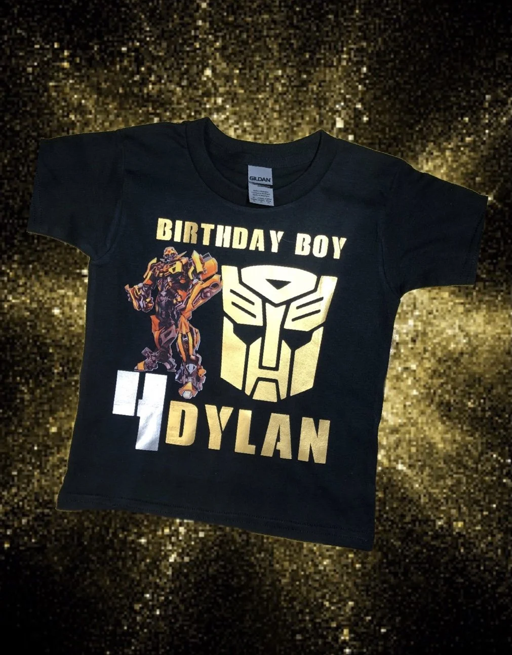 Black children's T-shirt with gold and white text and images, featuring the phrase 'Birthday Boy D'Ylan,' a transformer robot graphic, and a yellow Transformers Autobot symbol, set against a glittery gold background.