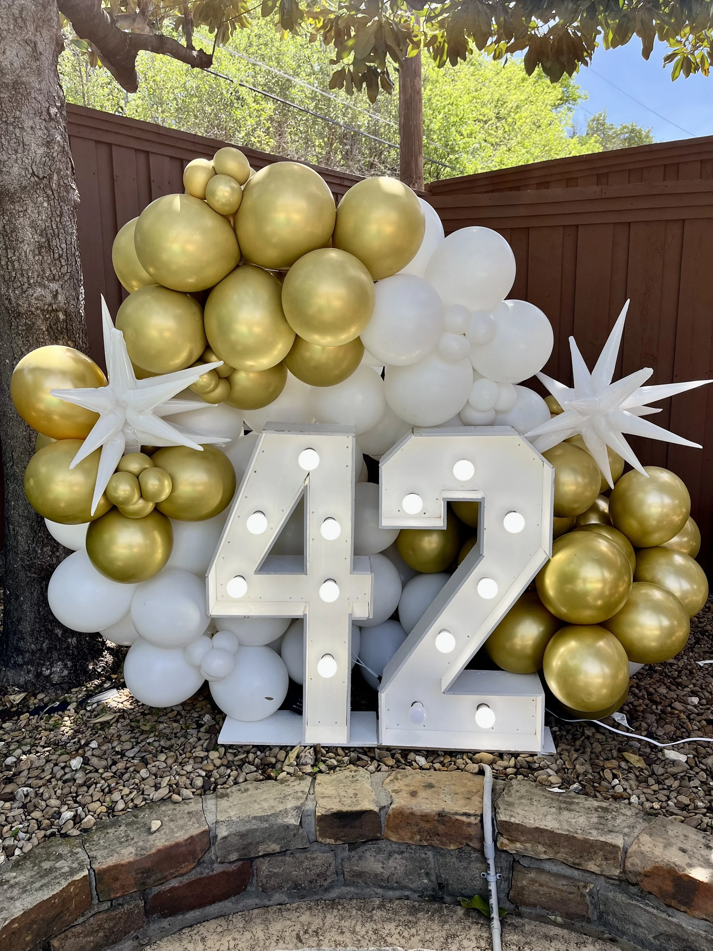 A decorative balloon arrangement featuring white and gold balloons, with star-shaped white balloons, set against a wooden fence and greenery on a sunny day. Large illuminated number '21' sign is in front of the balloons.