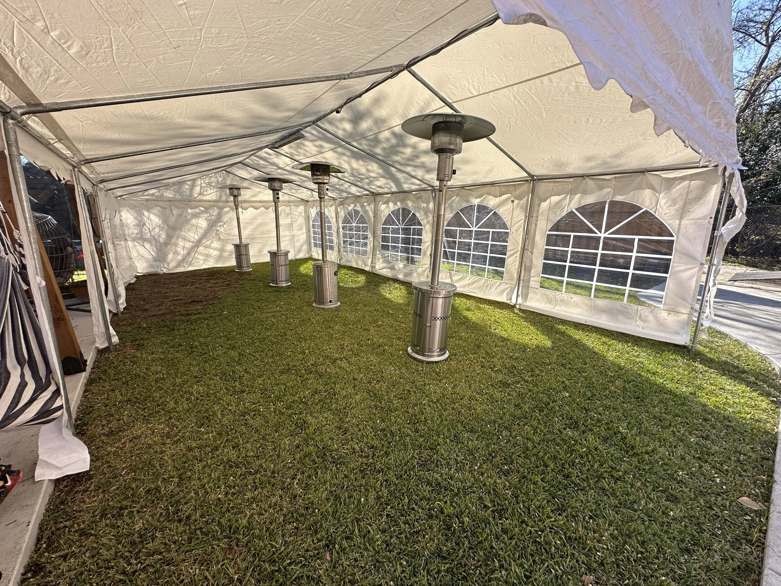 Inside of a white event tent with grass floor, electric patio heaters, and windows, on a sunny day.
