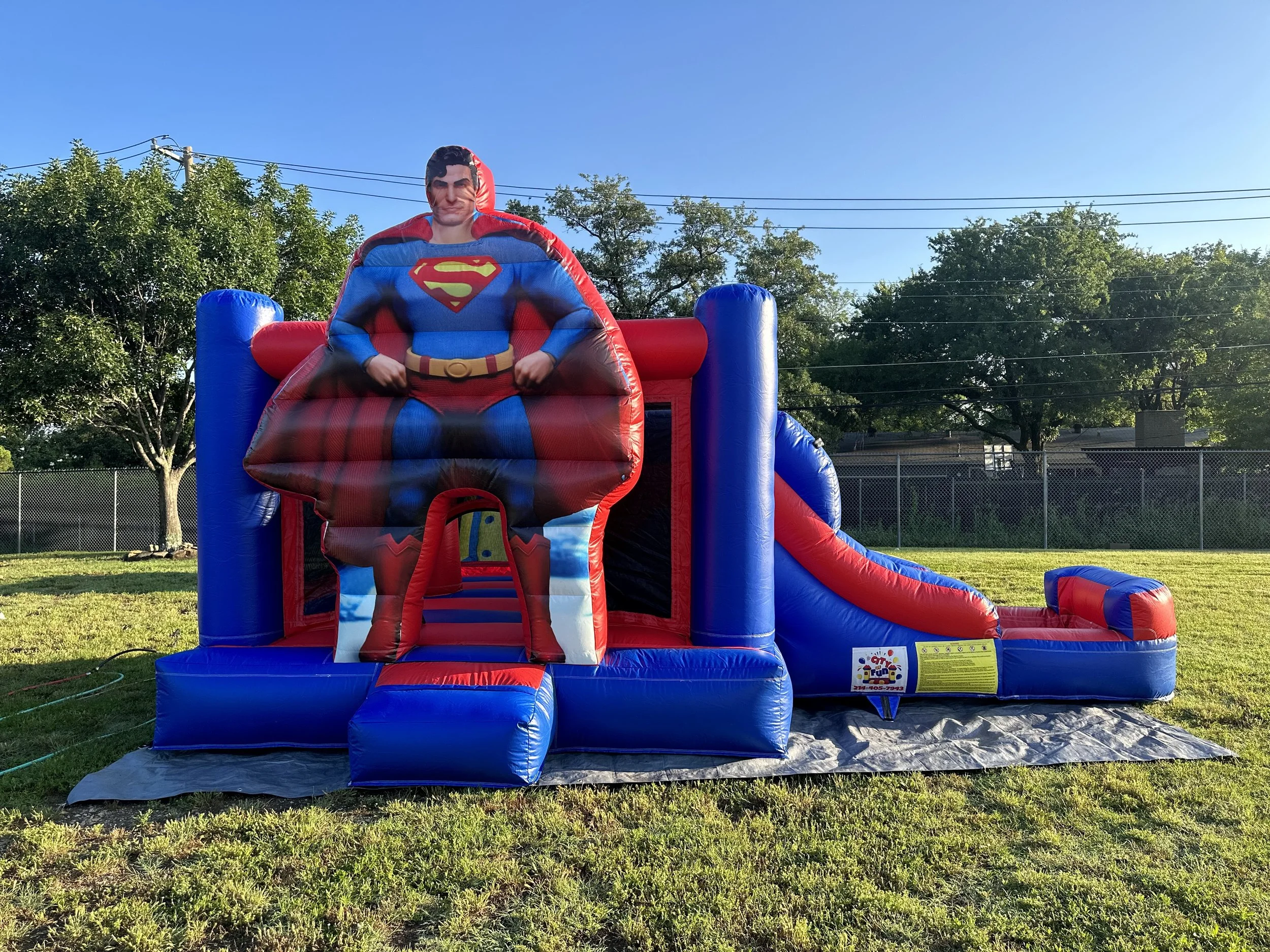 A large inflatable bounce house designed to look like Superman, with Superman's face and costume printed on the front. It is set up outdoors on a grassy area on a sunny day, with trees, a chain-link fence, and utility wires in the background.