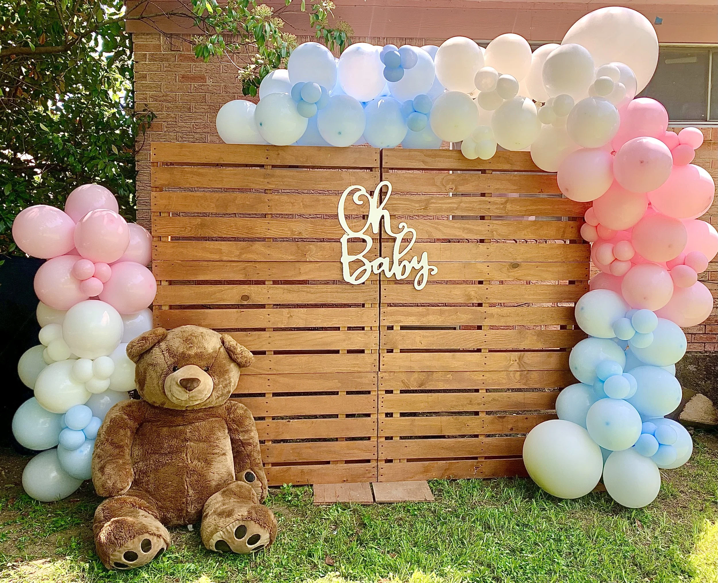 A wooden backdrop decorated with pastel-colored balloons in pink, blue, and white, with a sign that says 'Oh Baby' in cursive. A teddy bear is sitting on the grass in front of the backdrop.