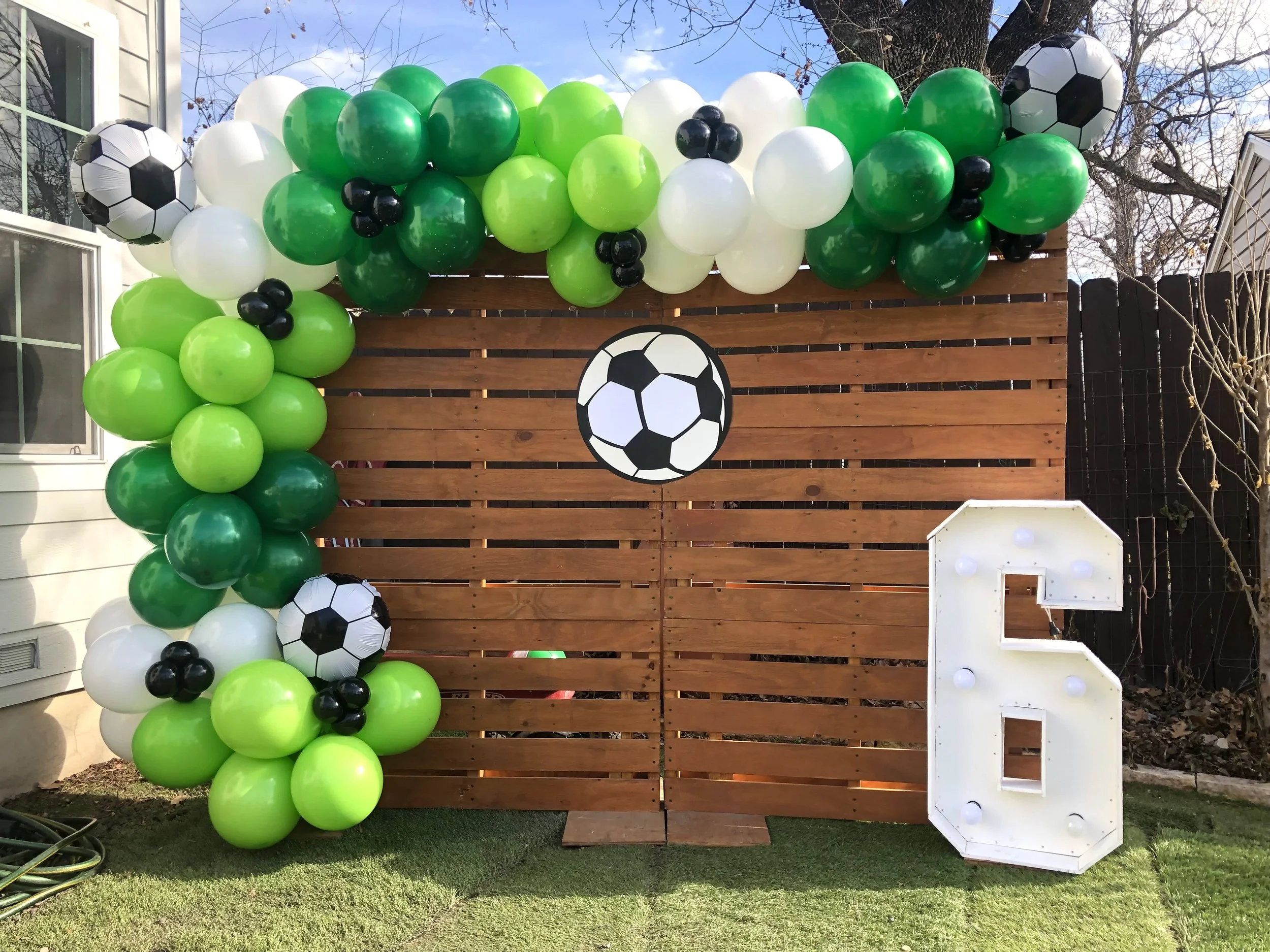 Decorative outdoor wall with green, white, and black balloons, soccer ball decorations, and a large white letter 'L' with lights.