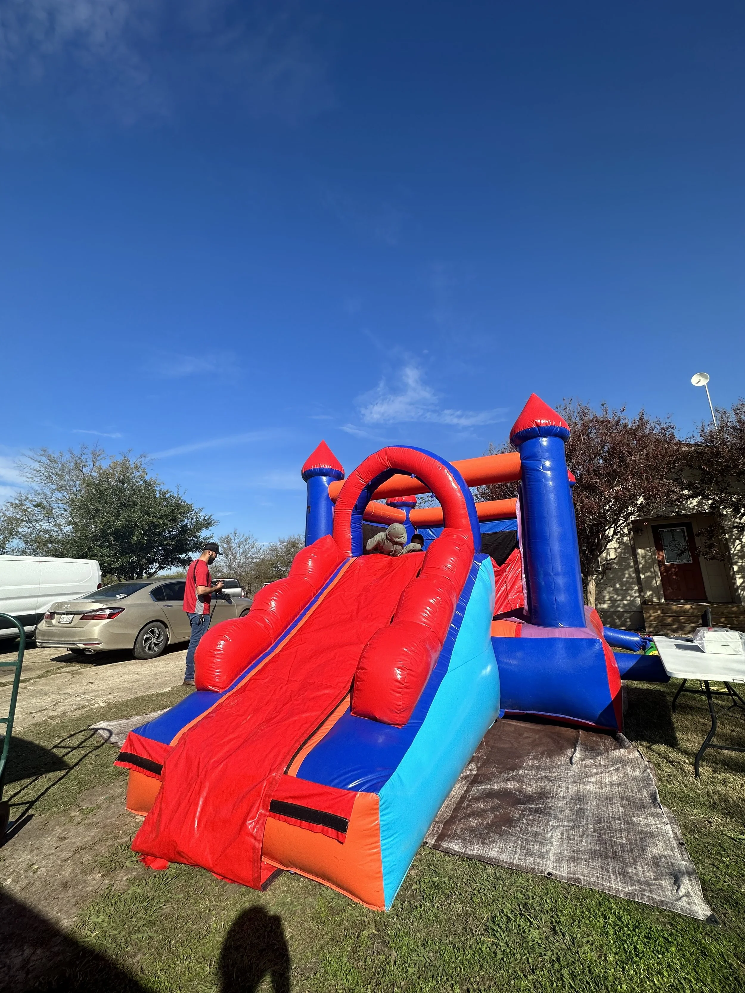 Colorful inflatable bounce house with a slide, set up outdoors in a parking lot area on a sunny day, with trees, cars, and a building in the background.