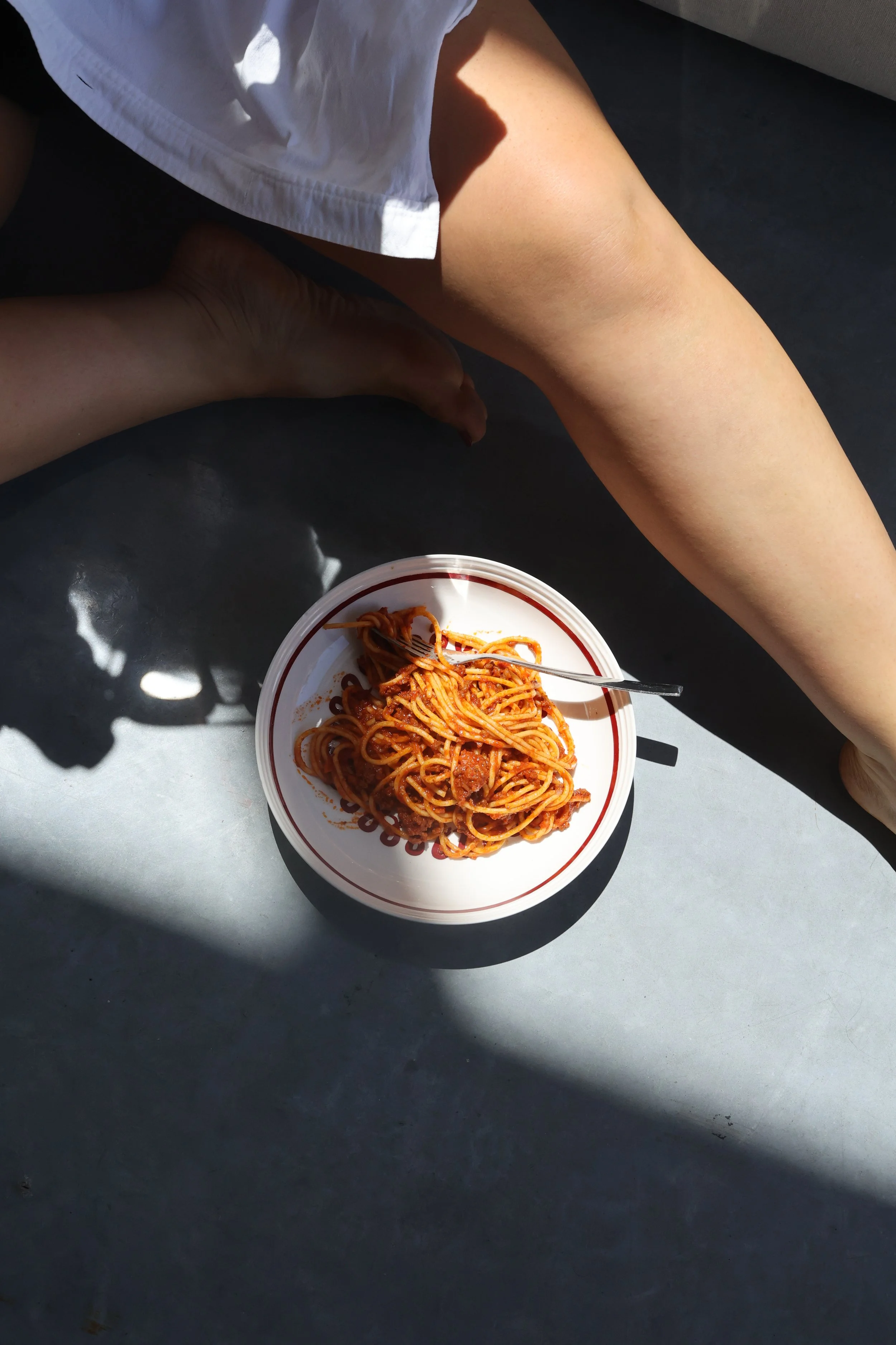 A bowl of spaghetti with meat sauce on a surface with a person's leg and foot nearby, illuminated by sunlight.