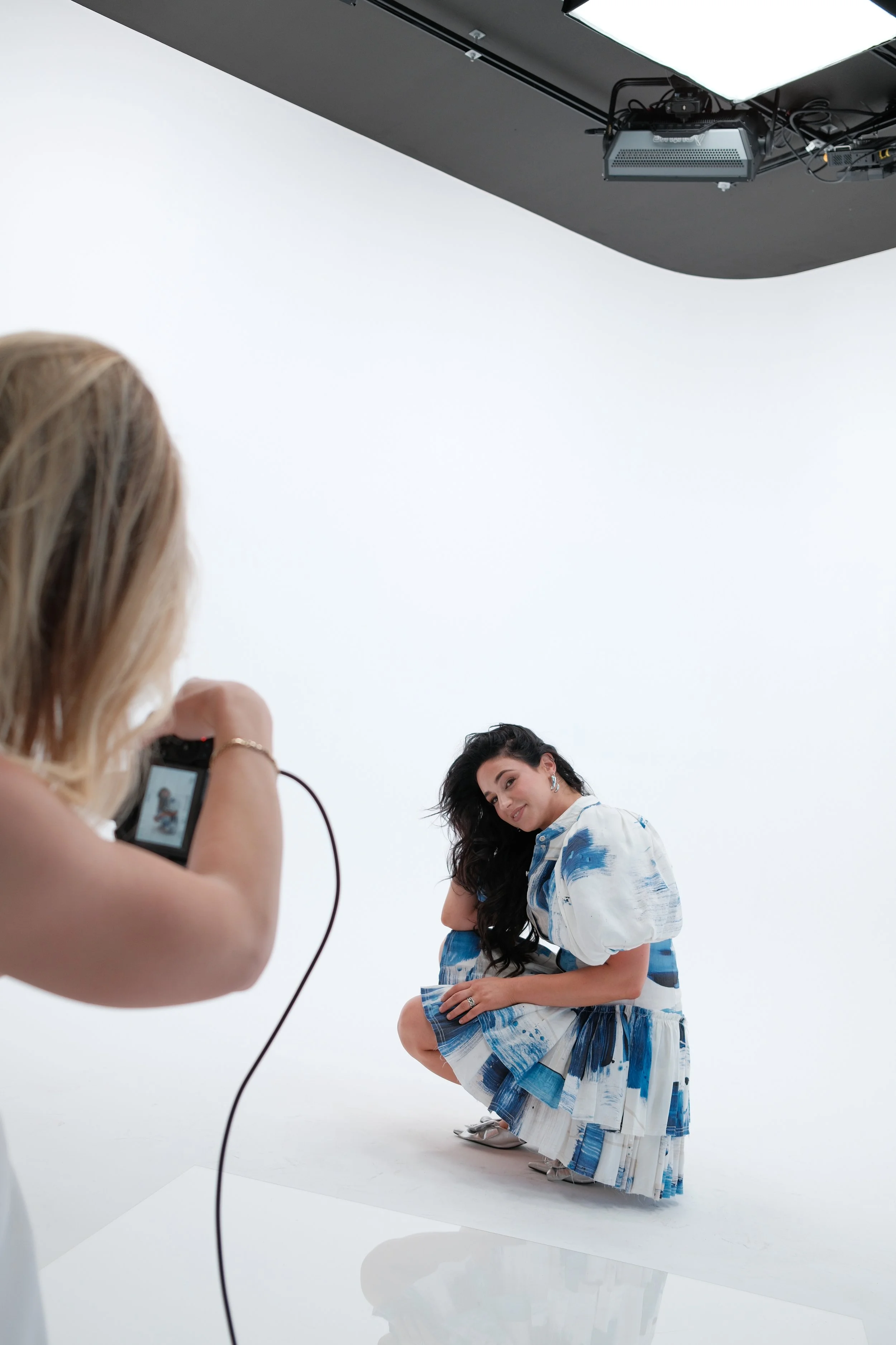 A woman with long black hair squatting on a white studio floor, wearing a white and blue patterned dress, while another woman with blonde hair takes her photo with a camera in a professional photography studio in Newcastle.