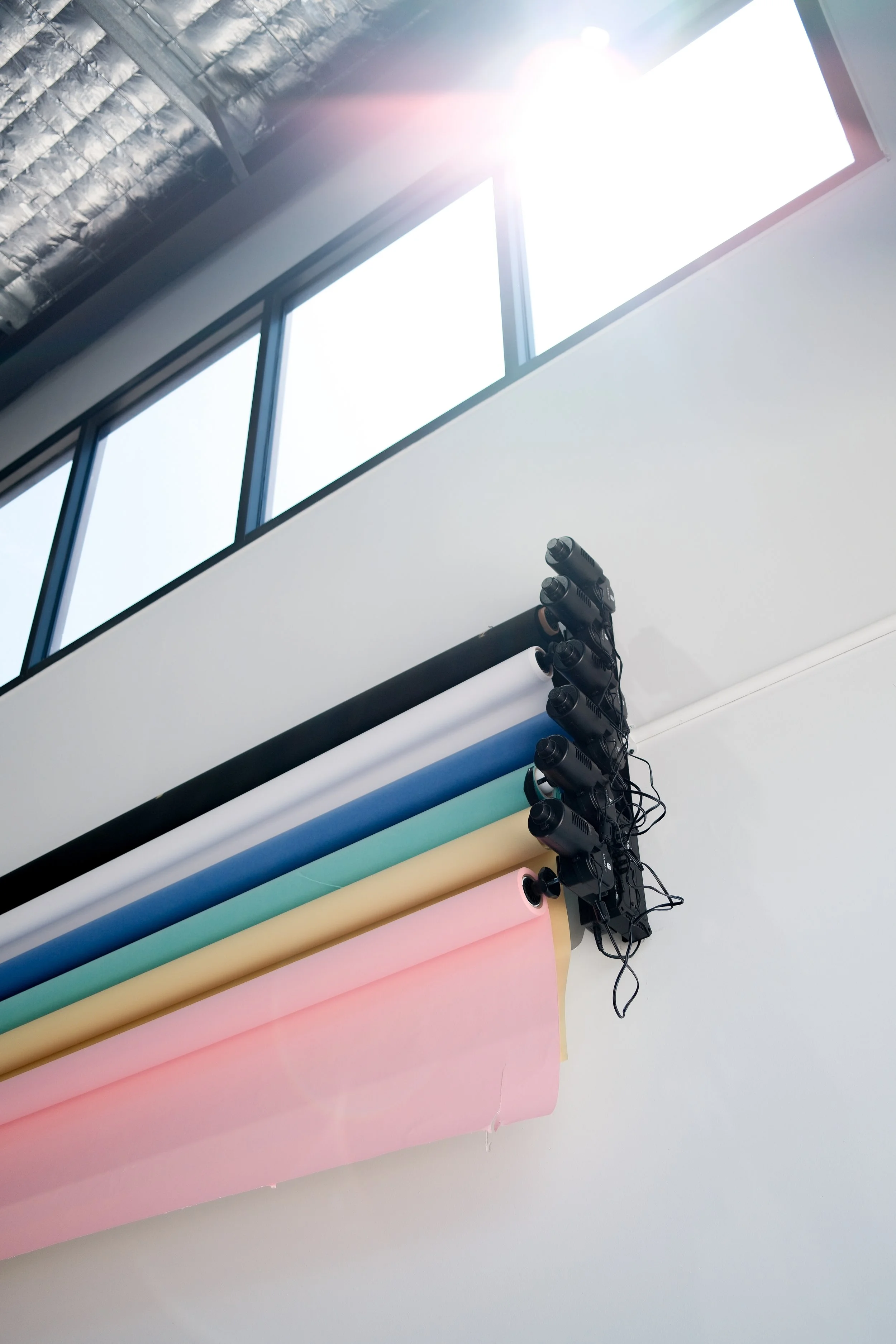 Colorful rolls of backdrop paper in pink, yellow, green, blue, white, and black hanging from a lighting rig in a photography studio with a large window overhead.