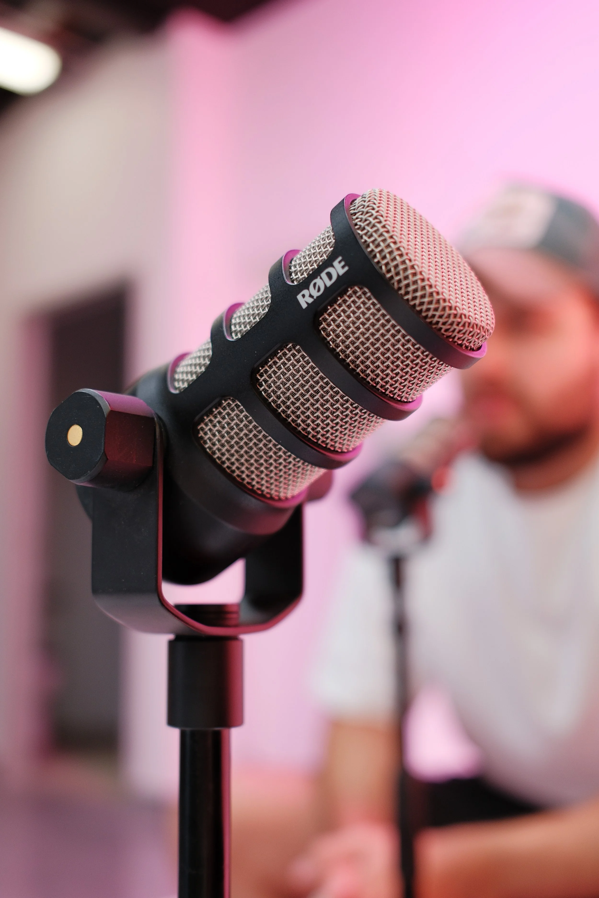 Close-up of a Rode microphone mounted on a stand, with a person in the background with a blurred face, set against a pink background.