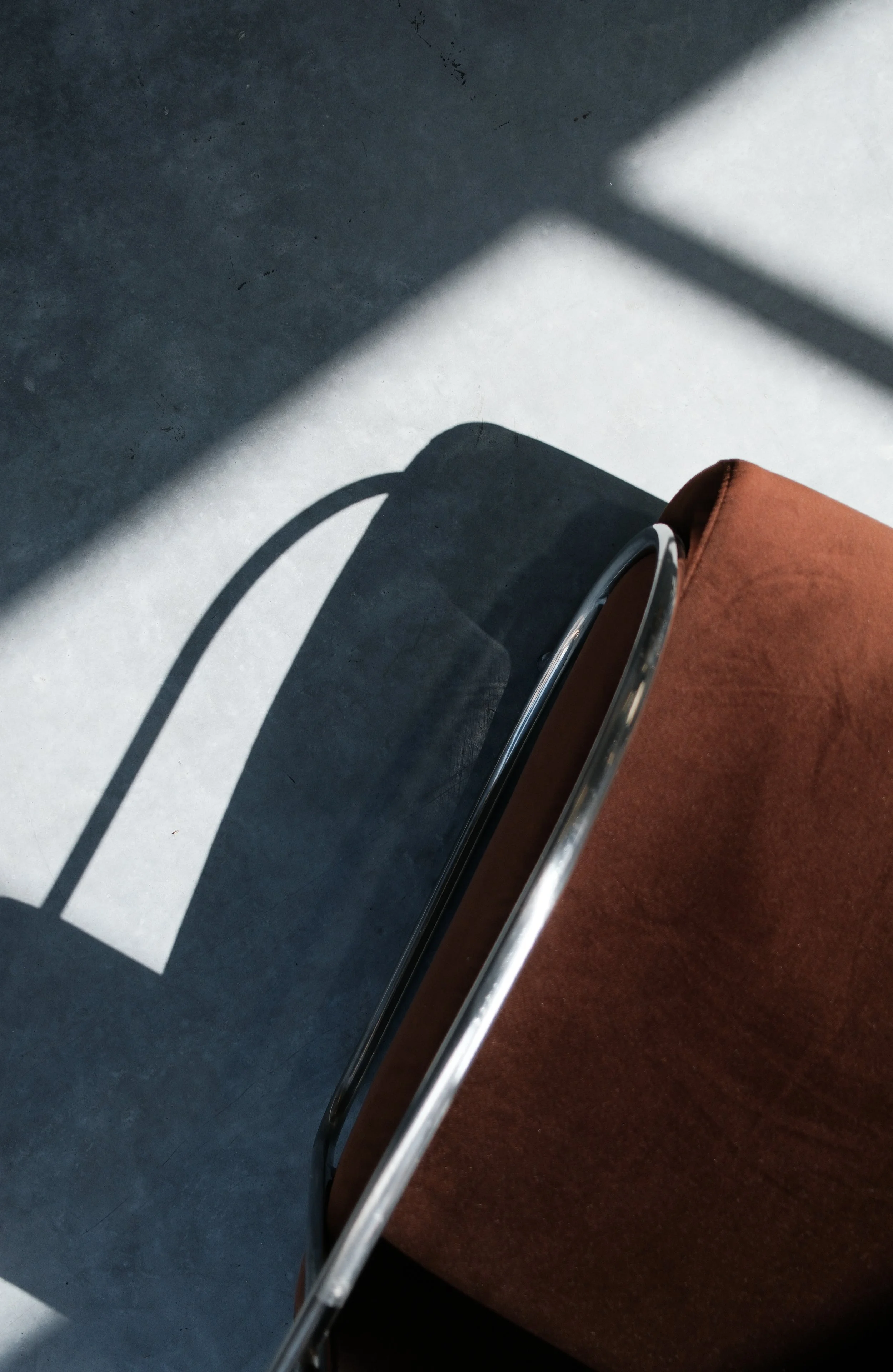 A brown armchair casting a shadow on a concrete floor with sunlight creating geometric patterns.