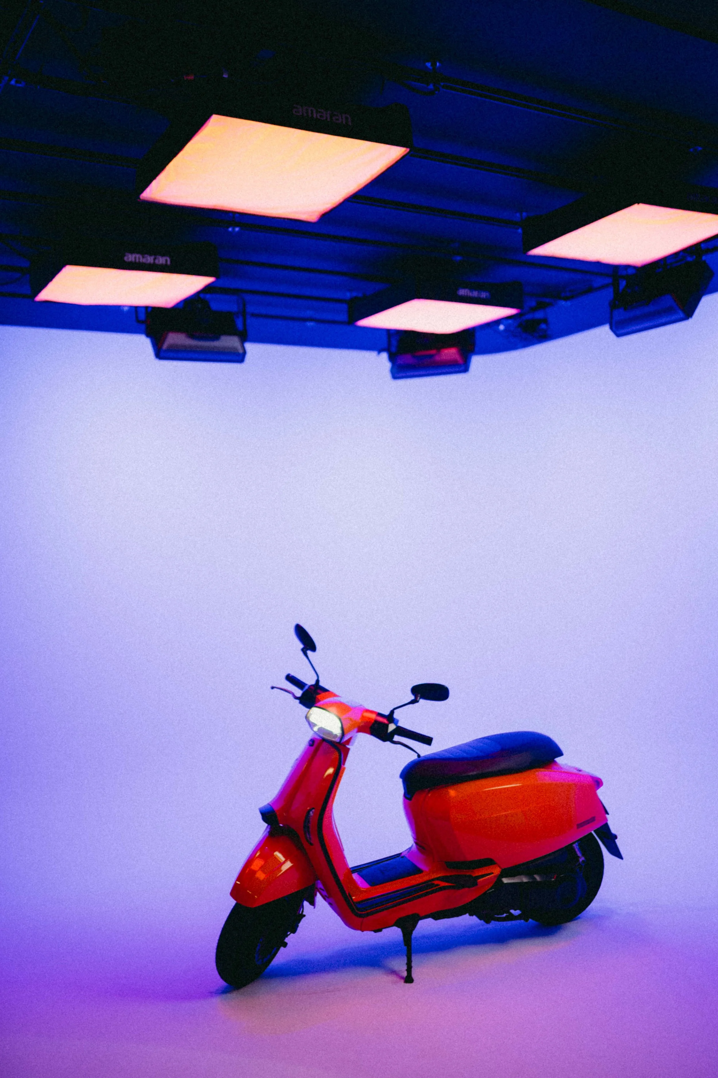 Orange scooter on a cyclorama with purple lighting, overhead lights are pink and purple, background is plain.