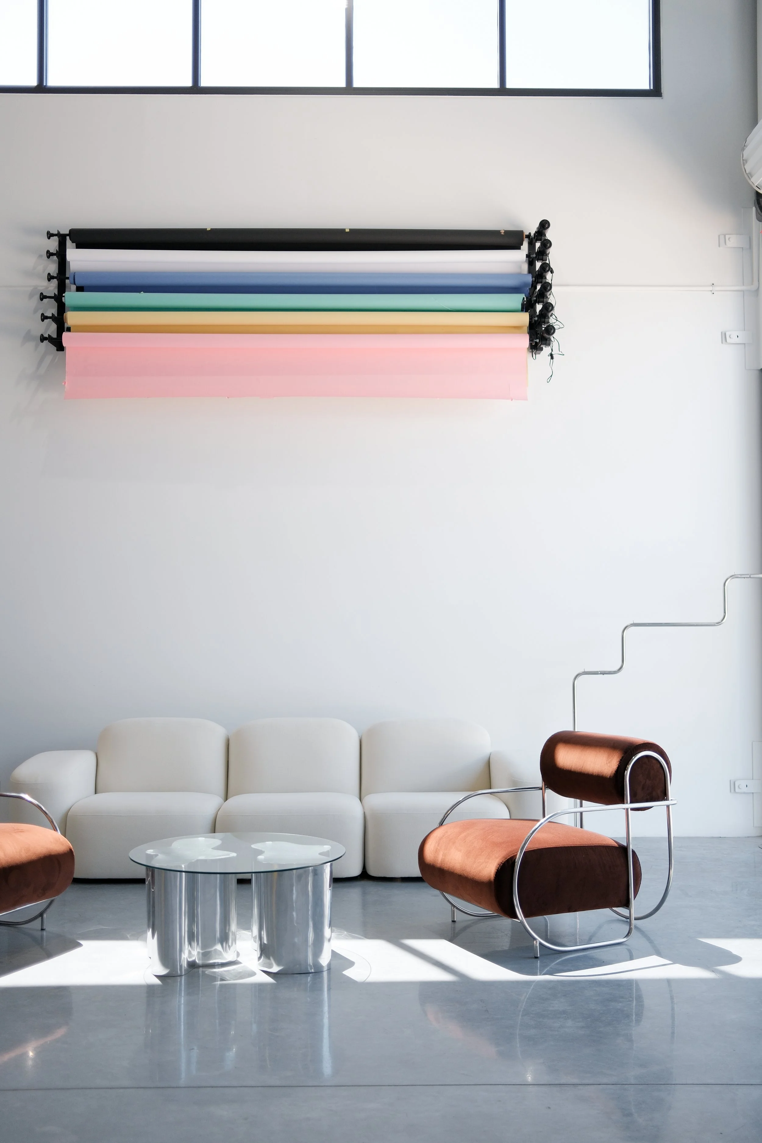 Minimalist modern natural light studio for hire in Newcastle with white sofa, dark brown armchairs, a glass coffee table, and colourful paper rolls mounted on the wall.