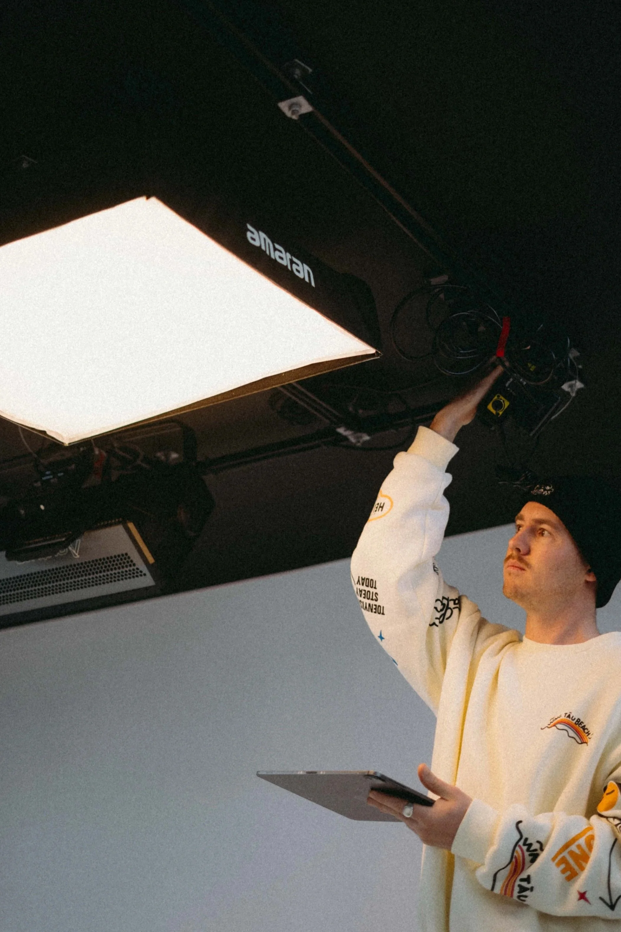 A man in a white sweatshirt and black beanie adjusting Amaran lighting equipment above a white cyclorama in Newcastle, NSW.
