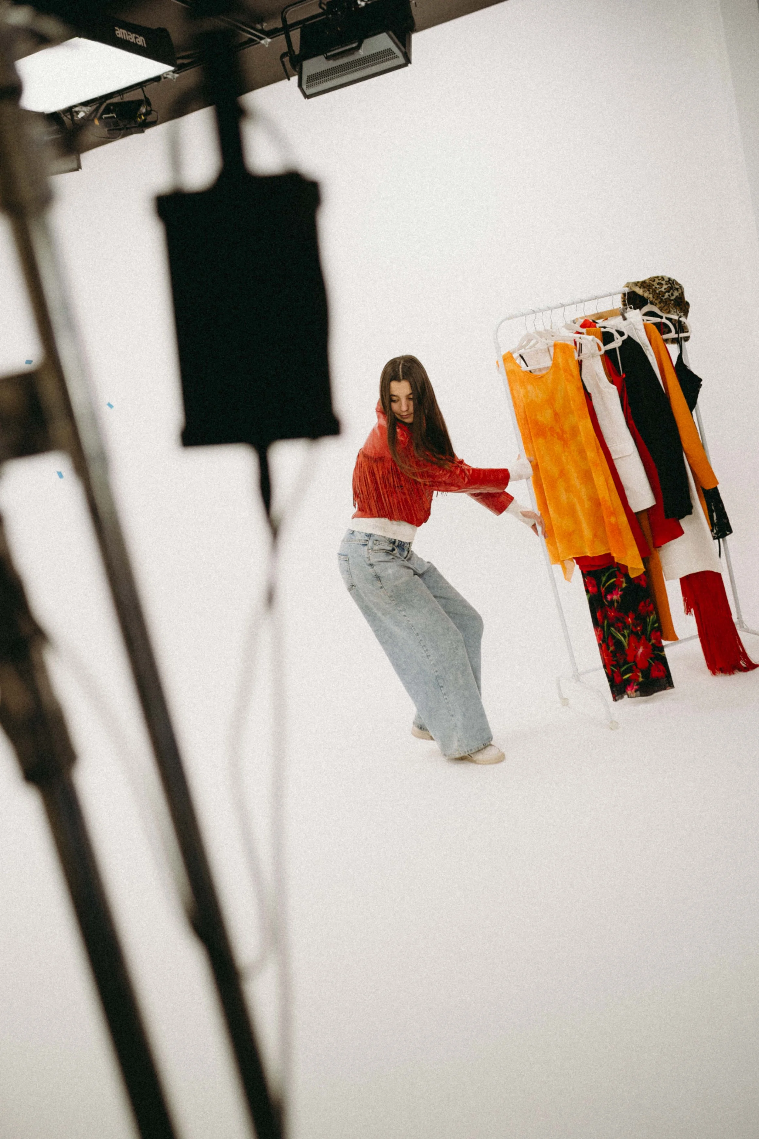 A woman with long brown hair dancing in a photo studio next to a clothing rack with colorful dresses and skirts, seen through studio lighting equipment.