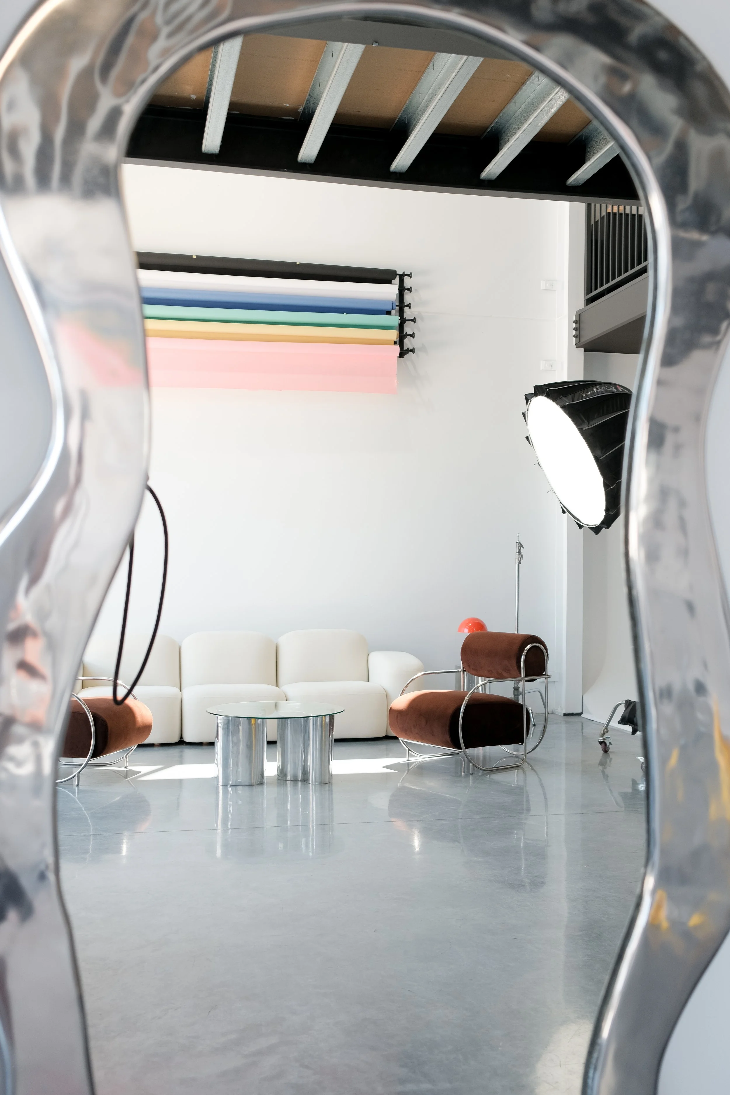 Modern interior space seen through a decorative metallic mirror, featuring a white sofa, brown and chrome chairs, a glass coffee table, colorful backdrop, studio lighting, and production equipment.