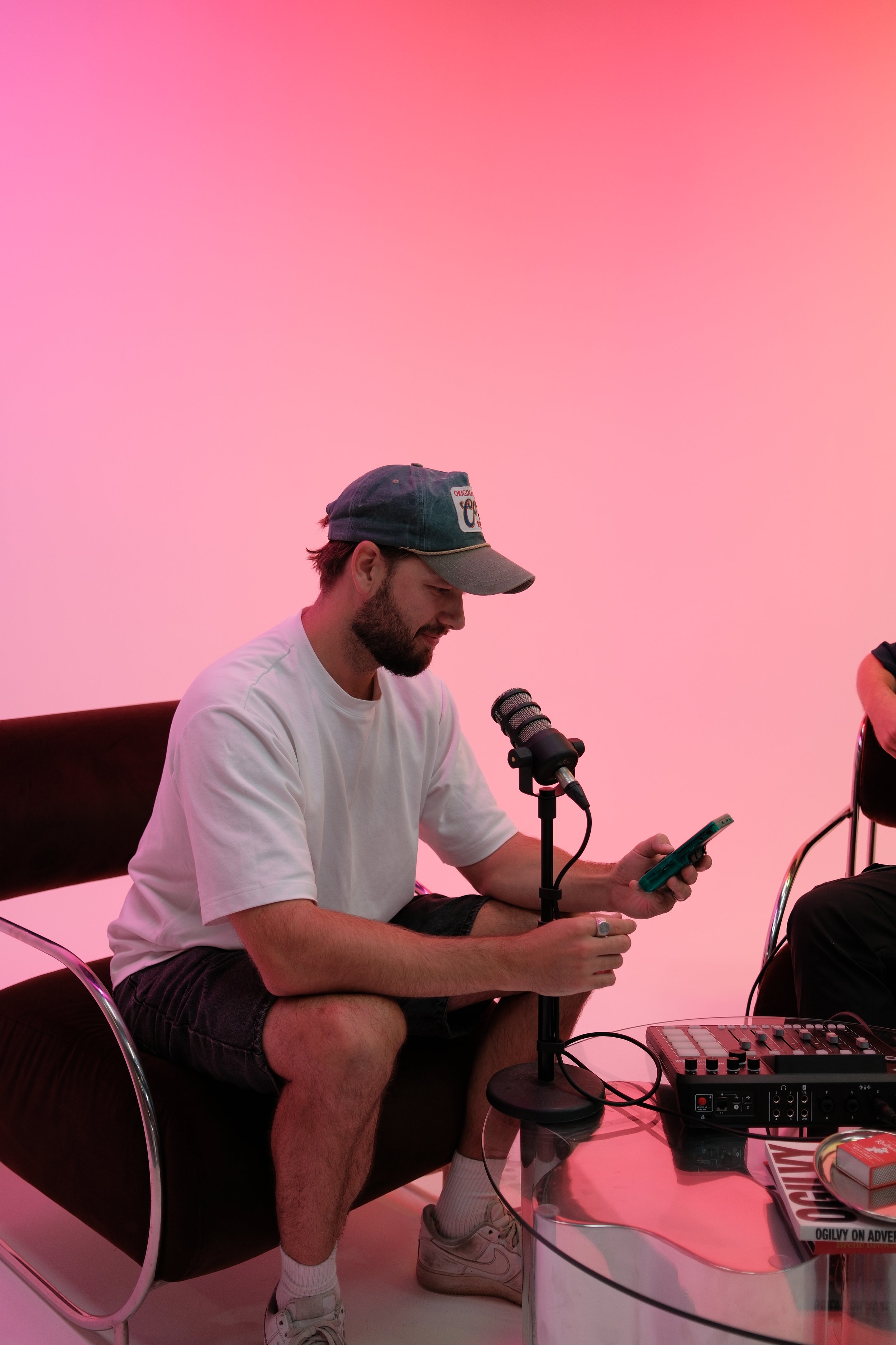 A man sitting on a black leather chair with a microphone in front of him, looking at his phone. He is wearing a white t-shirt, shorts, white socks, sneakers, and a baseball cap. There is audio equipment on the glass table next to him. The background 