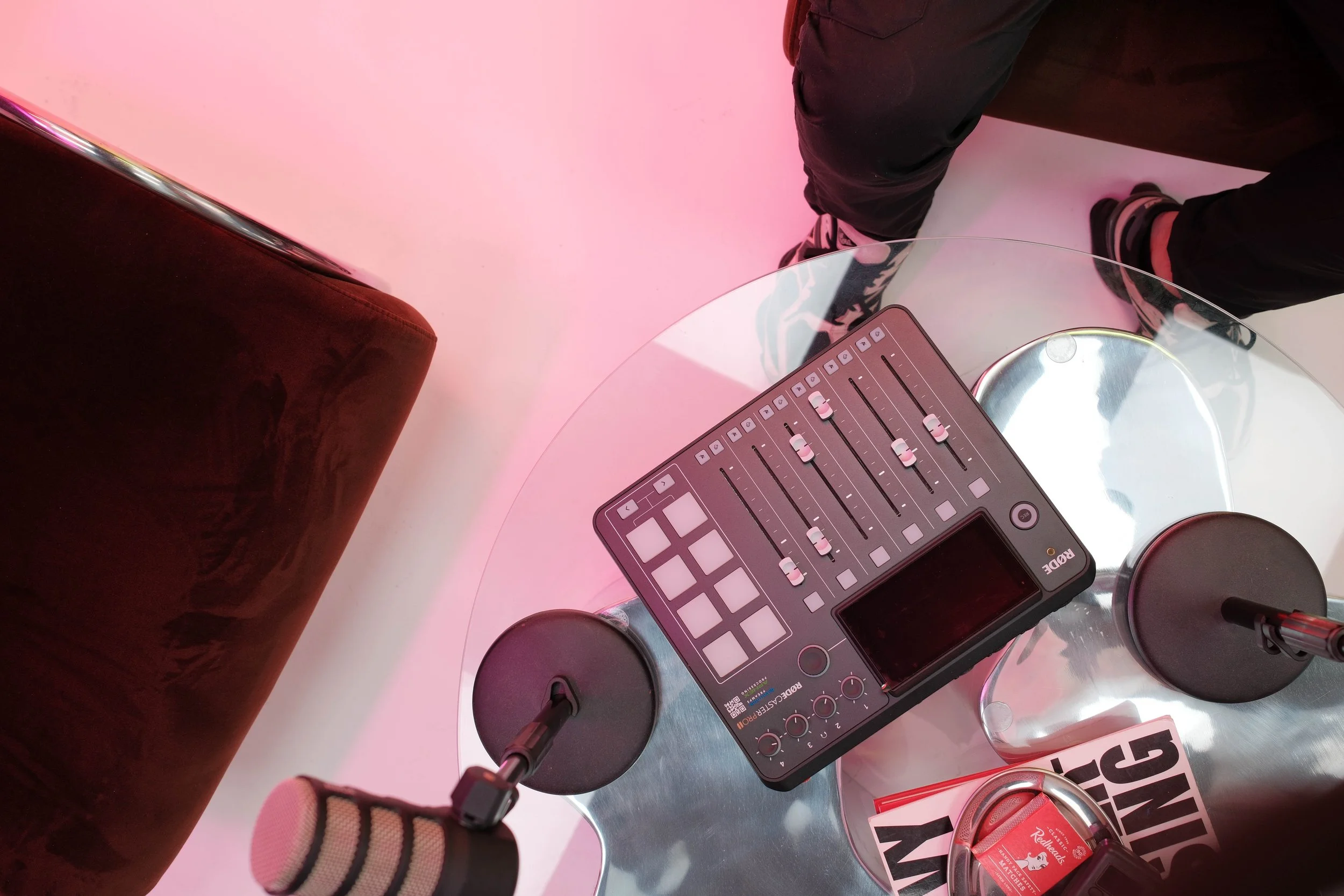 Top-down view of a podcast mixer placed on a glass table with microphones on either side, and of a person sitting nearby with legs in black pants and sneakers, in a space with pink lighting.