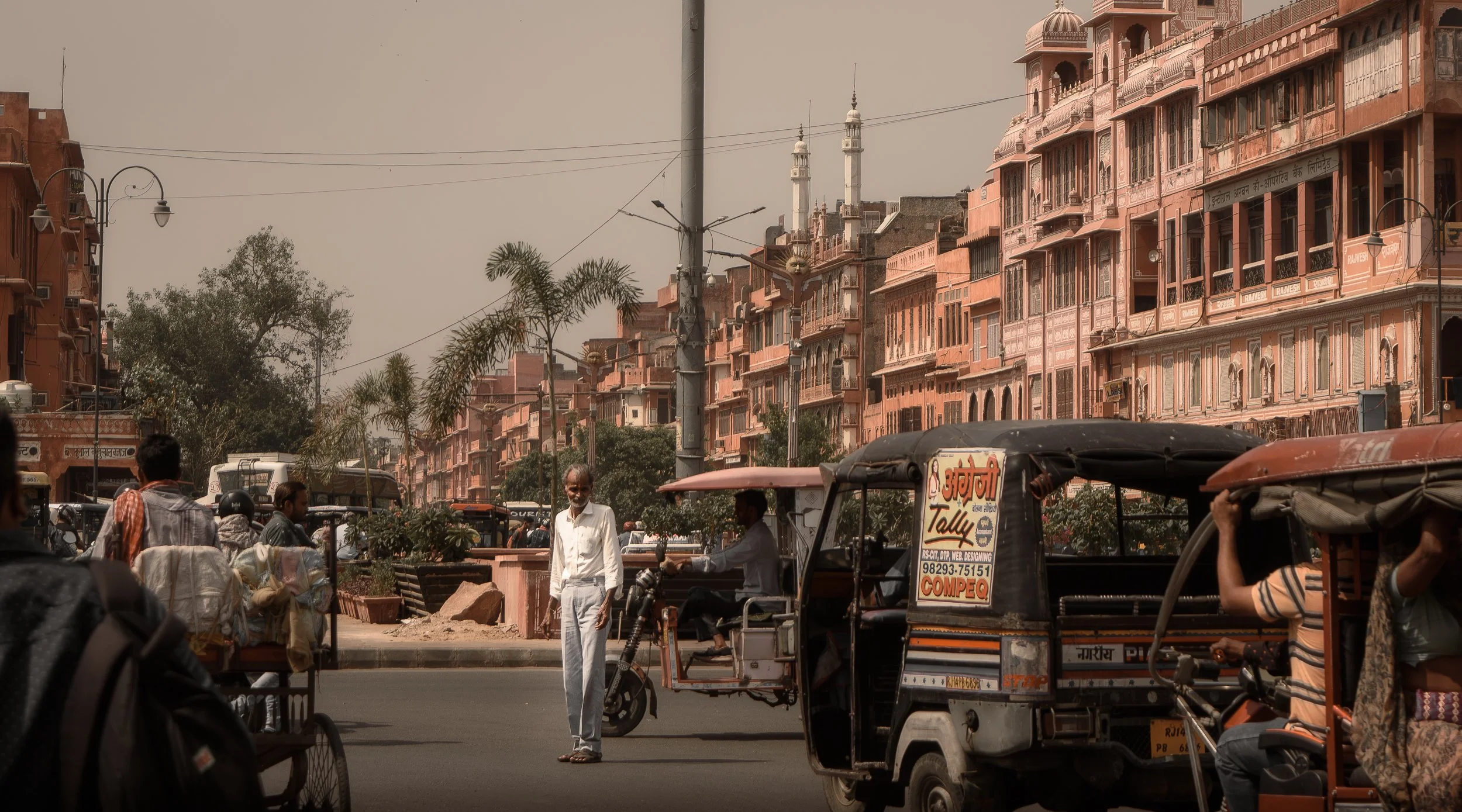 A street scene in Jaipur, India, showing pink-hued buildings, rickshaws, and pedestrians under warm daylight — capturing everyday life in the historic Pink City.