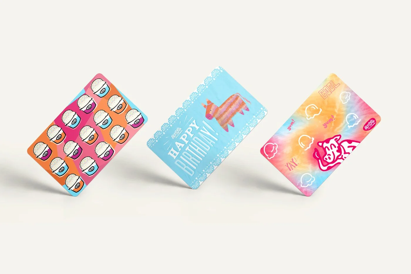 Row of 3 giftcards rotated to stand on their bottom left corner, illustrated collage of ice cream scoops in Amy's brand colors, colorful illustrated piñata that reads Happy Birthday, tie-dye background with Yum, Cool, Yay floating around dancing cow