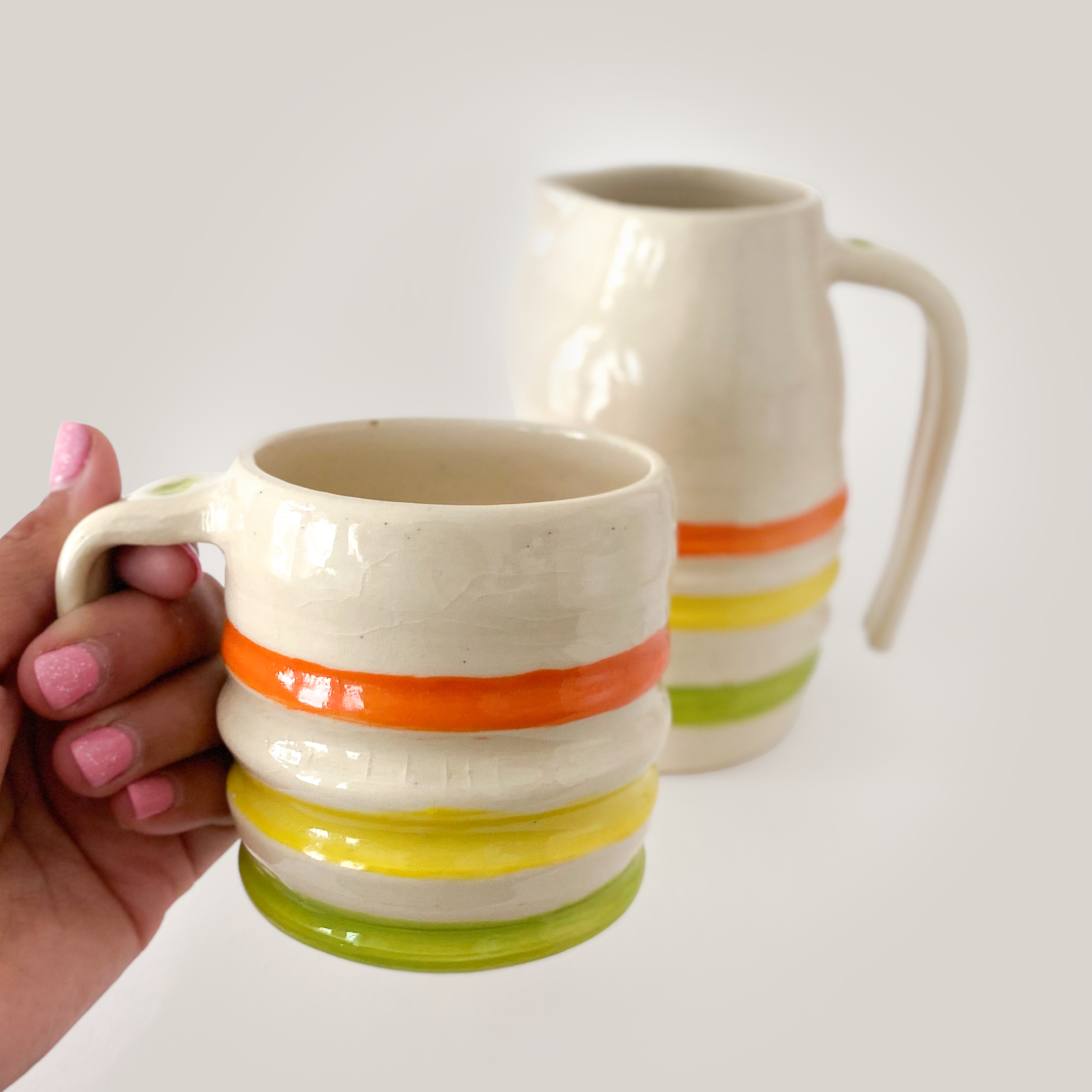 hand holding a mug with ridges painted in a orange, yellow, green pattern. Behind sits a pitcher with the same design.