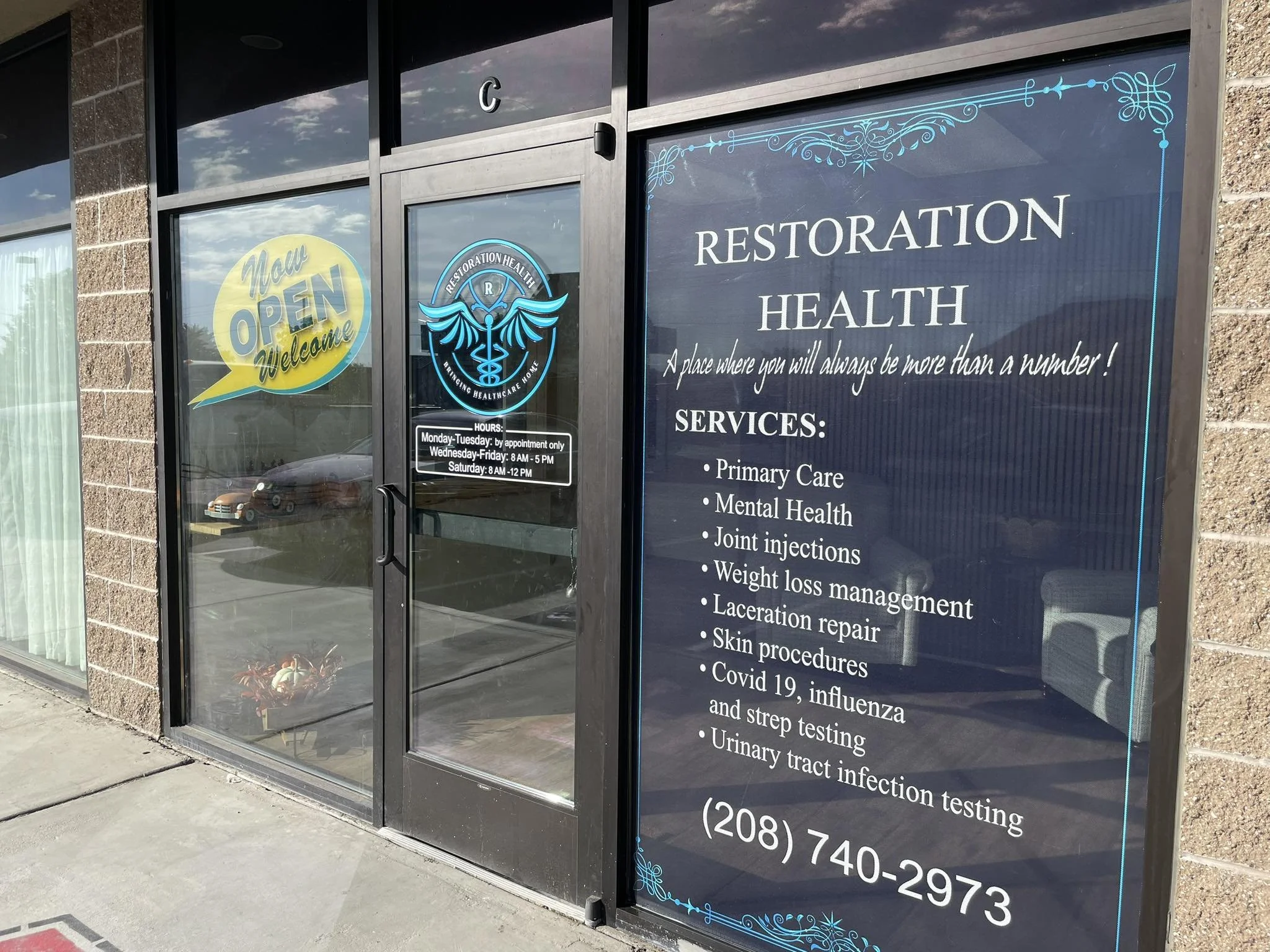 Glass doors and window of a healthcare clinic with yellow and blue signs indicating the clinic is now open and welcoming, and a large blue sign listing services such as primary care, mental health, joint injections, weight management, skin procedures, infectious disease testing, and urinary tract infections, along with the clinic's phone number.