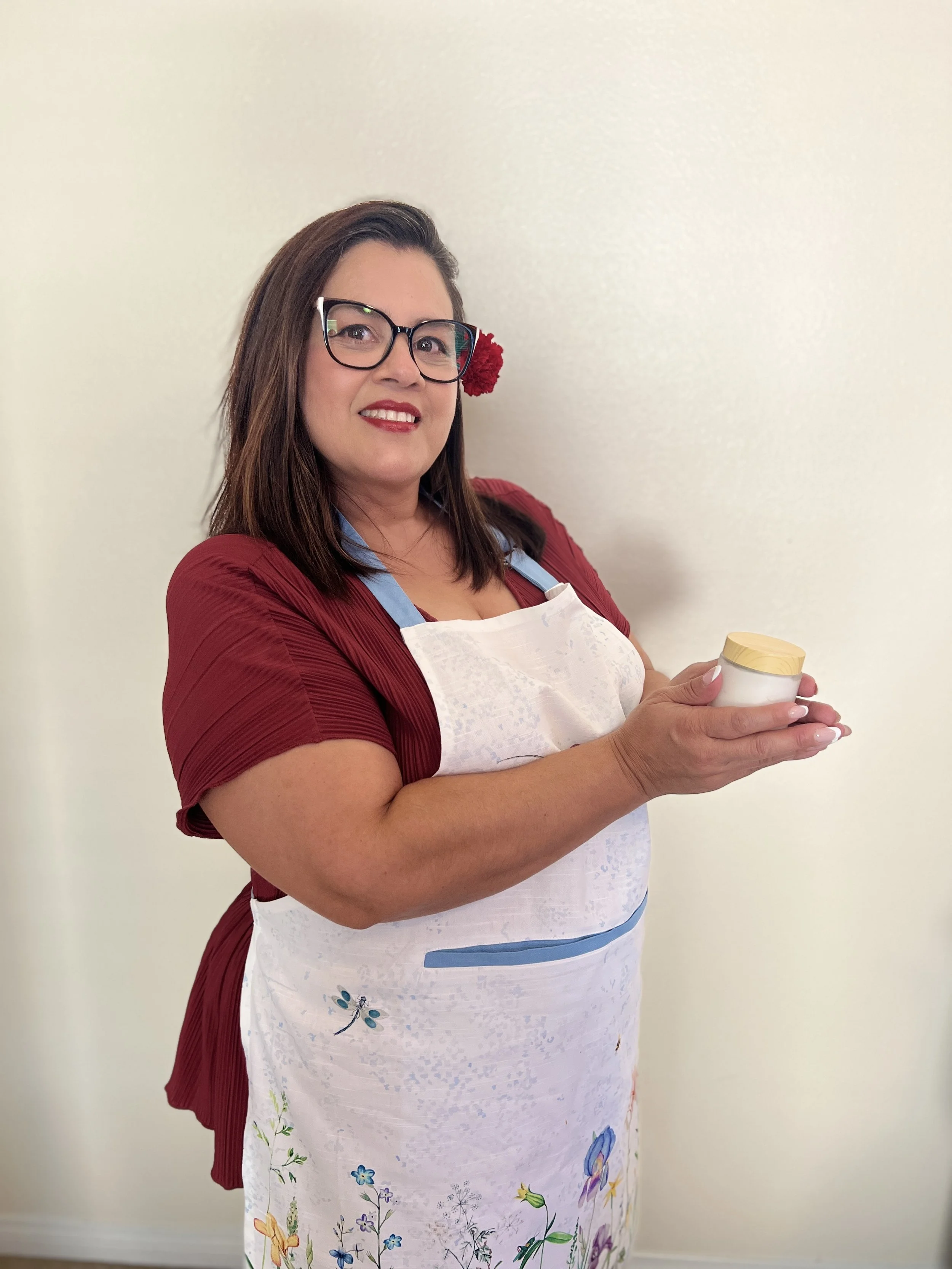 A woman with glasses and a red flower in her hair, wearing a red dress and a white apron with floral and butterfly designs, holding a container, standing against a plain light-colored wall.