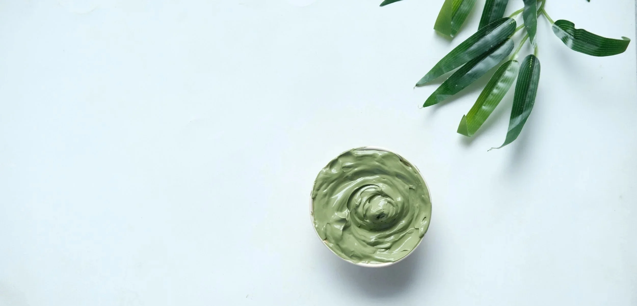 A white surface with green cream or paint swirled in a circle and some green leaves positioned in the top right corner.