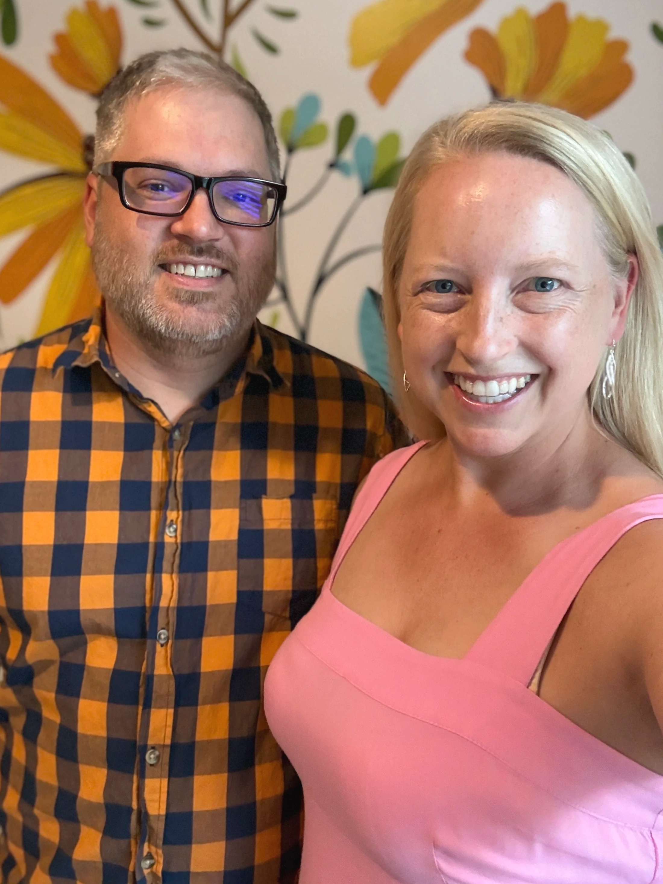 A man with glasses and a man with blonde hair smiling, standing close together in front of a colorful floral-patterned background.