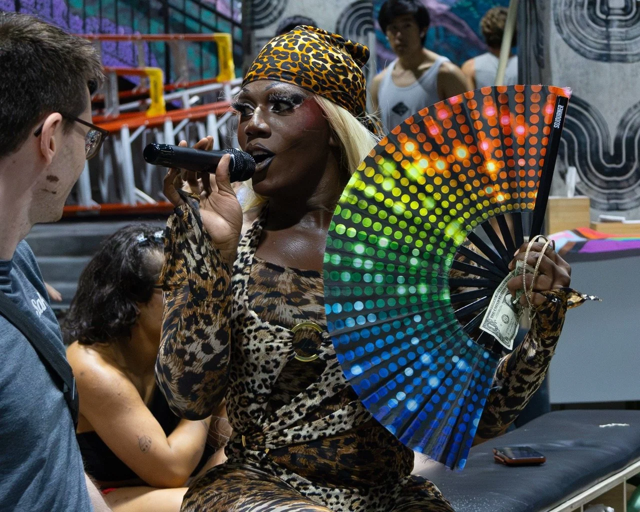 A black drag queen at a microphone speaking at a pride event in Washington DC holding a rainbow fan