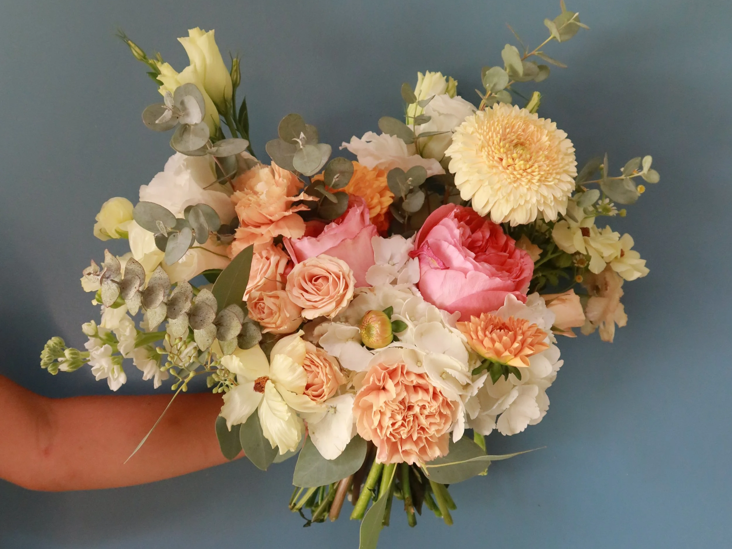A hand holds a small bouquet of mixed flowers including roses, gerbera daisies, lisianthus, eucalyptus, and other greenery against a blue background.