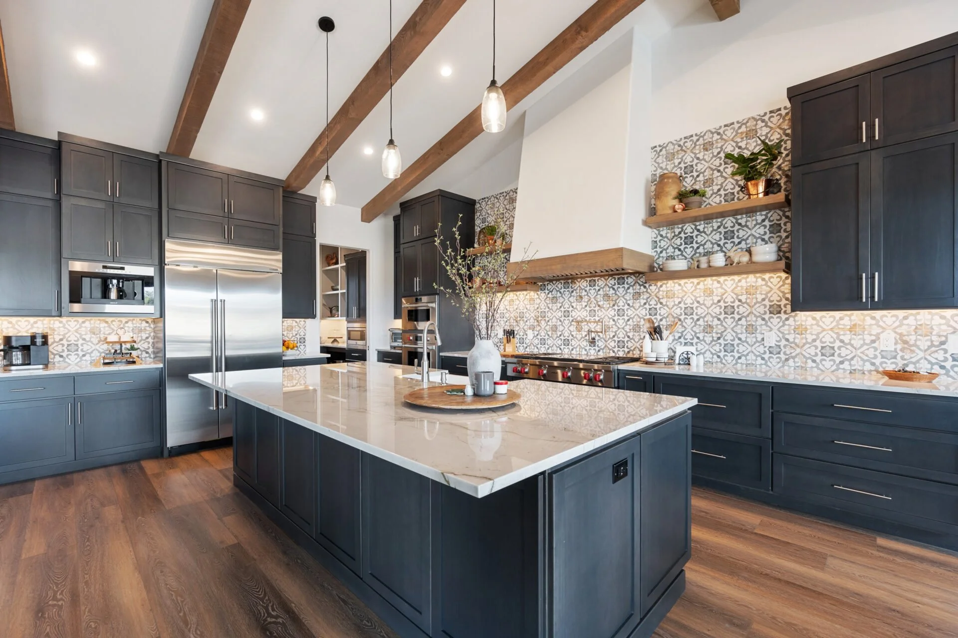 Modern kitchen with dark blue cabinets, a large island with a white marble countertop, wood beams on the ceiling, patterned backsplash tiles, open wooden shelves with plants and dishes, stainless steel appliances, and wooden flooring.