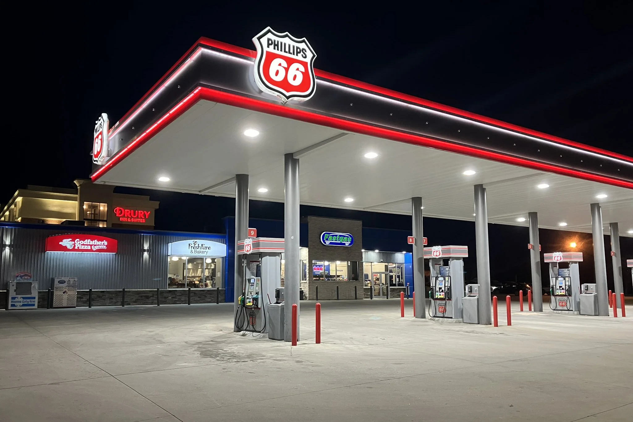 Night scene of a Phillips 66 gas station with multiple fuel pumps, illuminated canopy, and surrounding convenience stores, including Godfather's Pizza and Fresh Fare Bakery.