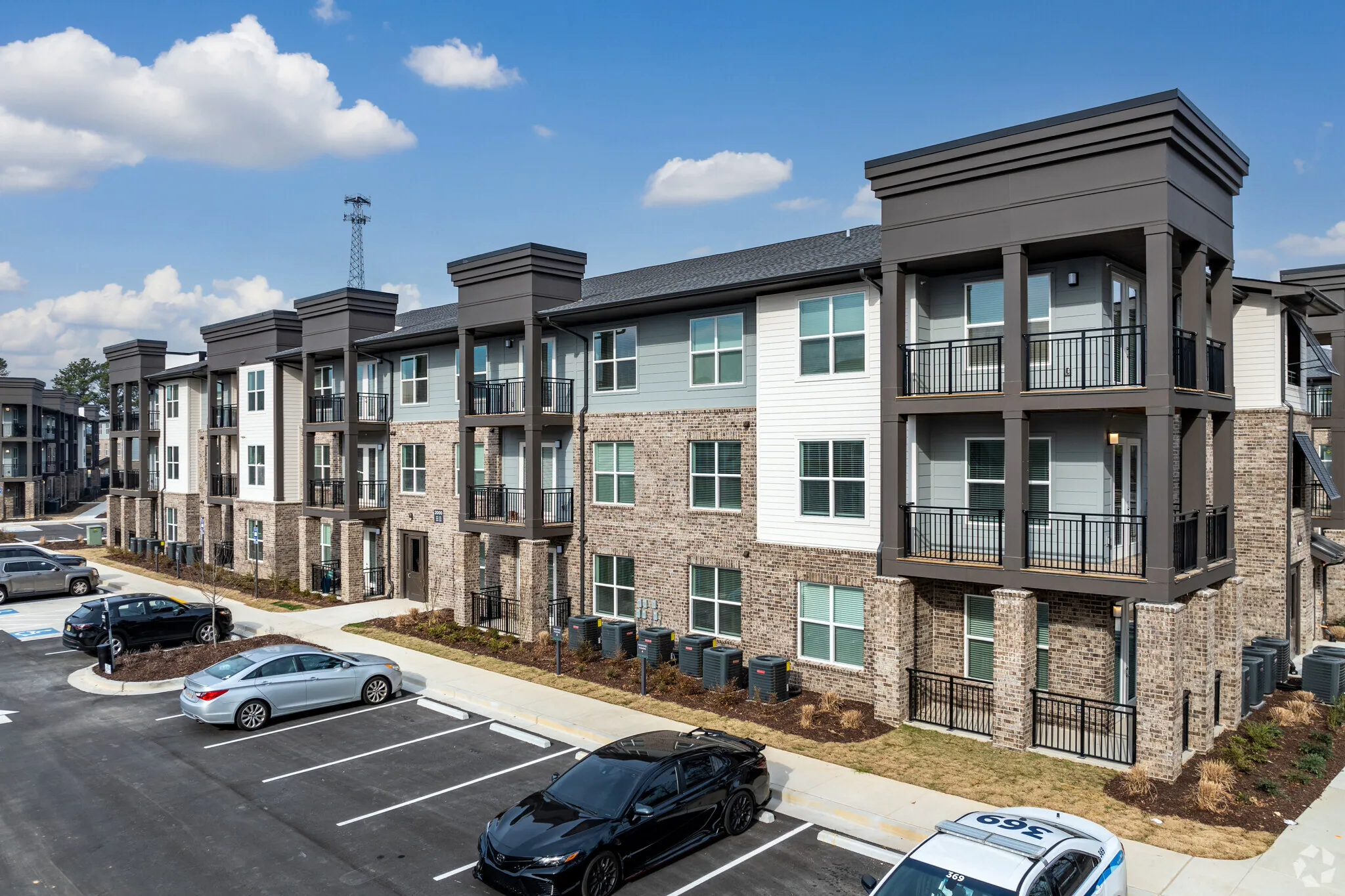 Exterior view of a modern apartment complex with brick and siding facades, balconies, and a parking lot with several parked cars under a partly cloudy sky.