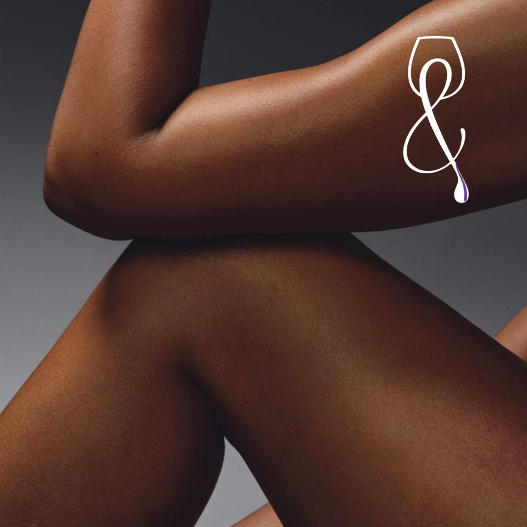 Close-up of a person's arm, torso, and thigh with smooth, dark skin against a gray background, featuring a white stylized ampersand symbol.