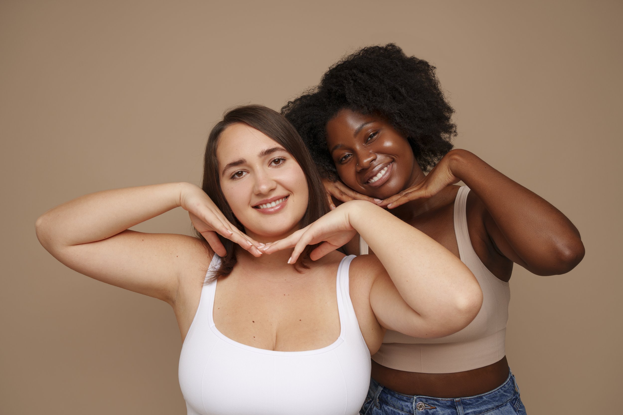 Two women smiling with their hands under their chins, wearing tank tops, standing against a plain beige background.