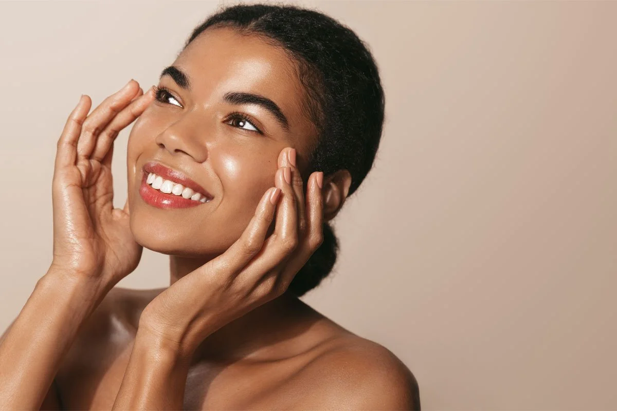 A woman with dark hair in a bun smiling and touching her face with her hands, against a neutral background.