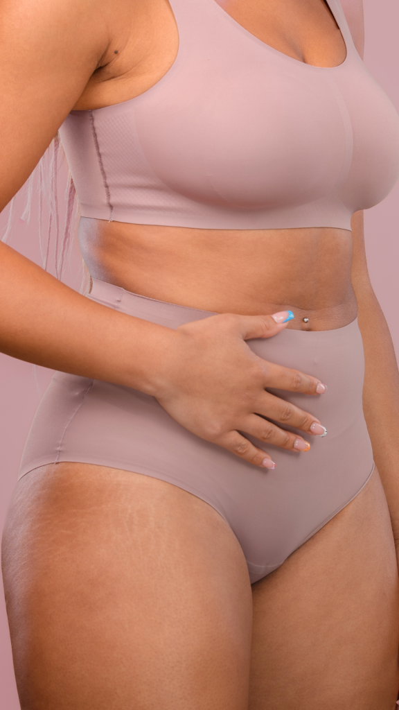 Close-up of a woman wearing nude-colored sports bra and high-waisted underwear, with her hand resting on her stomach and a belly button piercing.