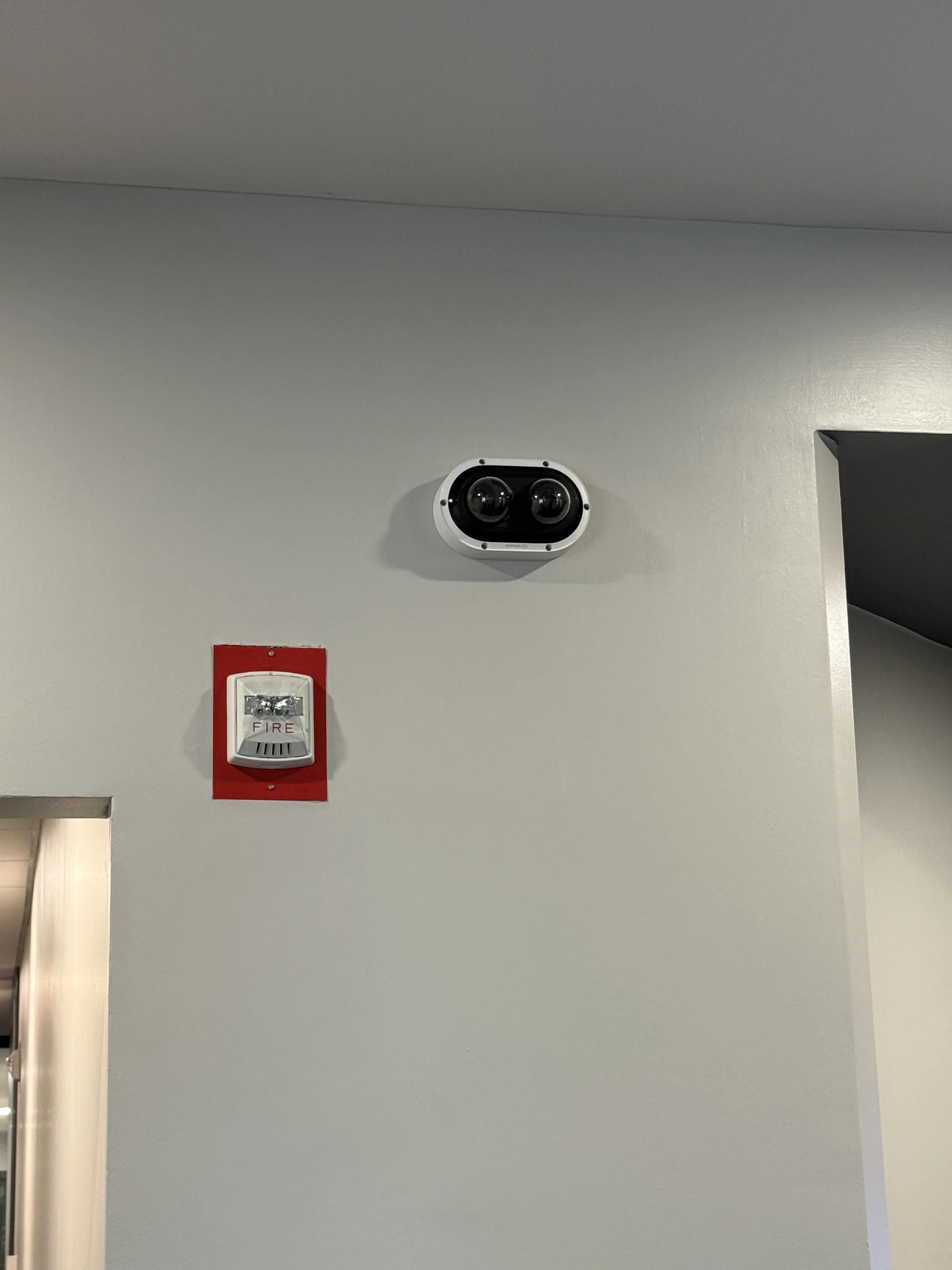 Security camera with two lenses mounted on a light gray wall above a fire alarm.