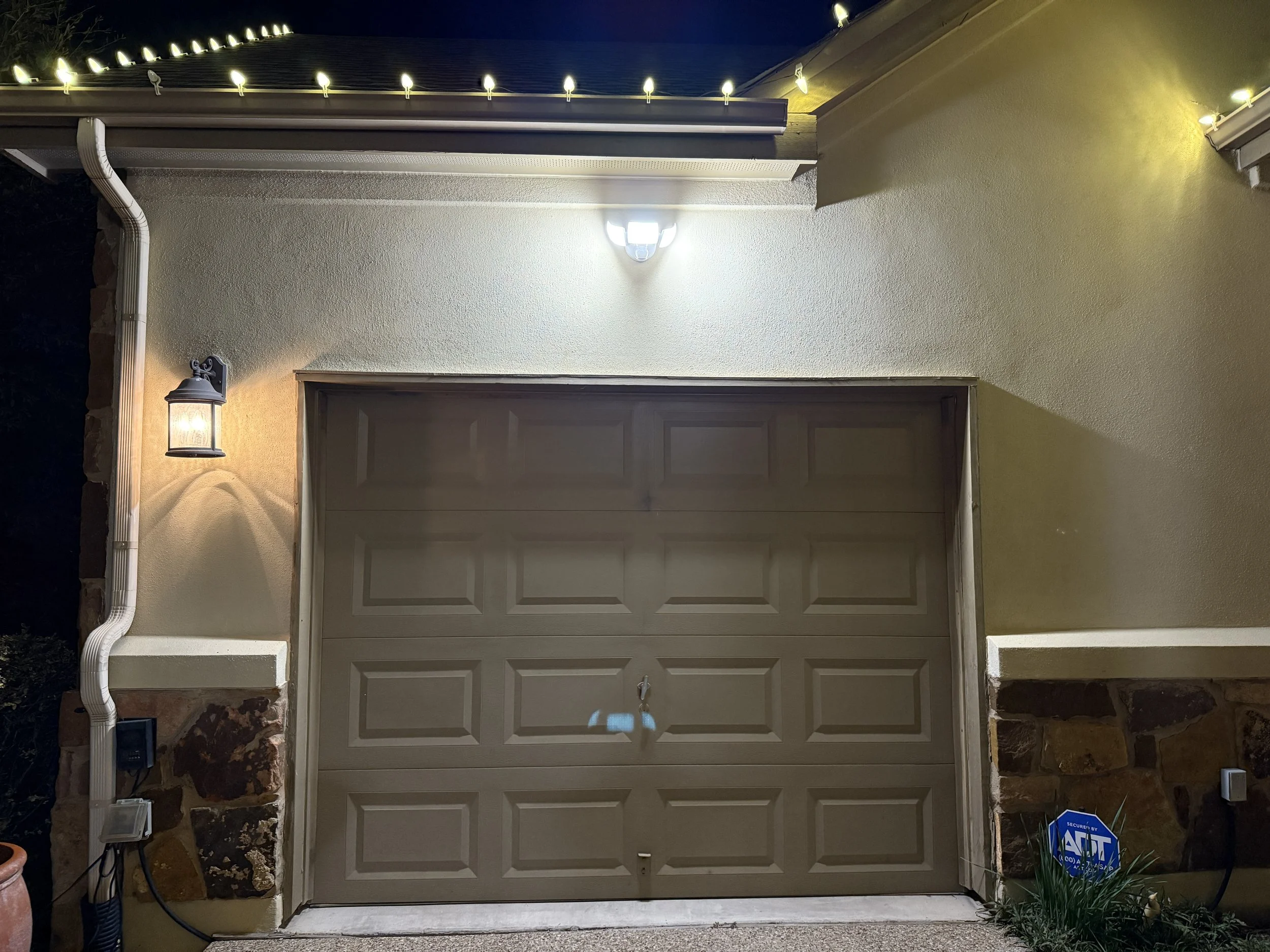 Exterior view of a garage door with porch lights and security alarm sign, illuminated at night.