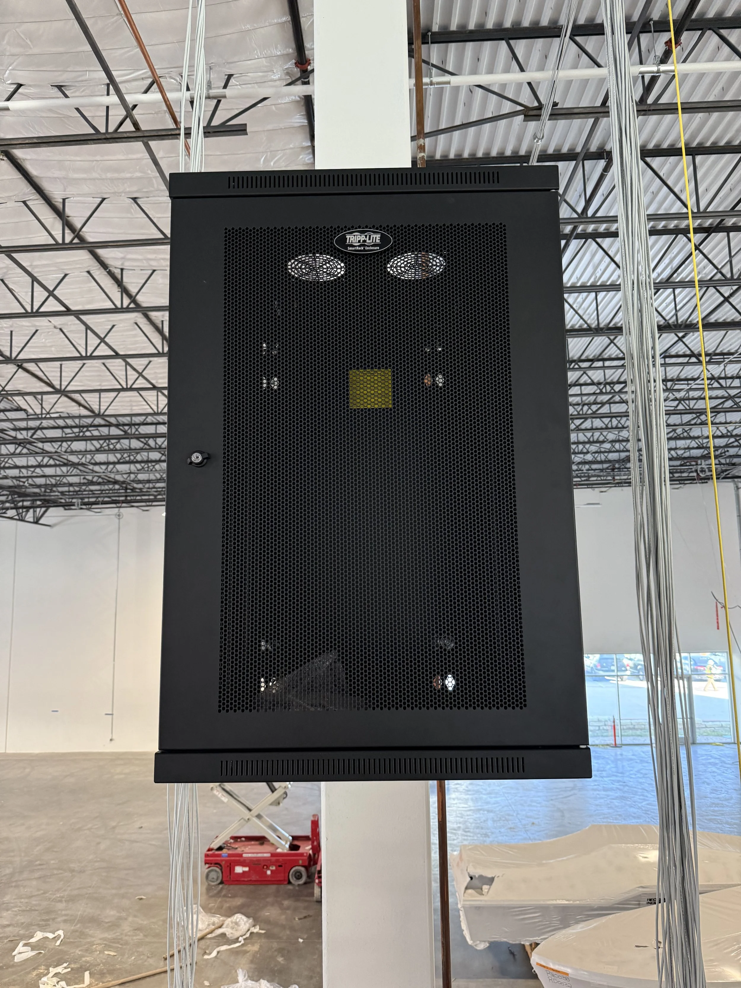 Black electrical or electronic equipment mounted on a wall in a large industrial space, with a metal grid front panel and ventilation holes, surrounded by construction materials and incomplete ceiling framework.