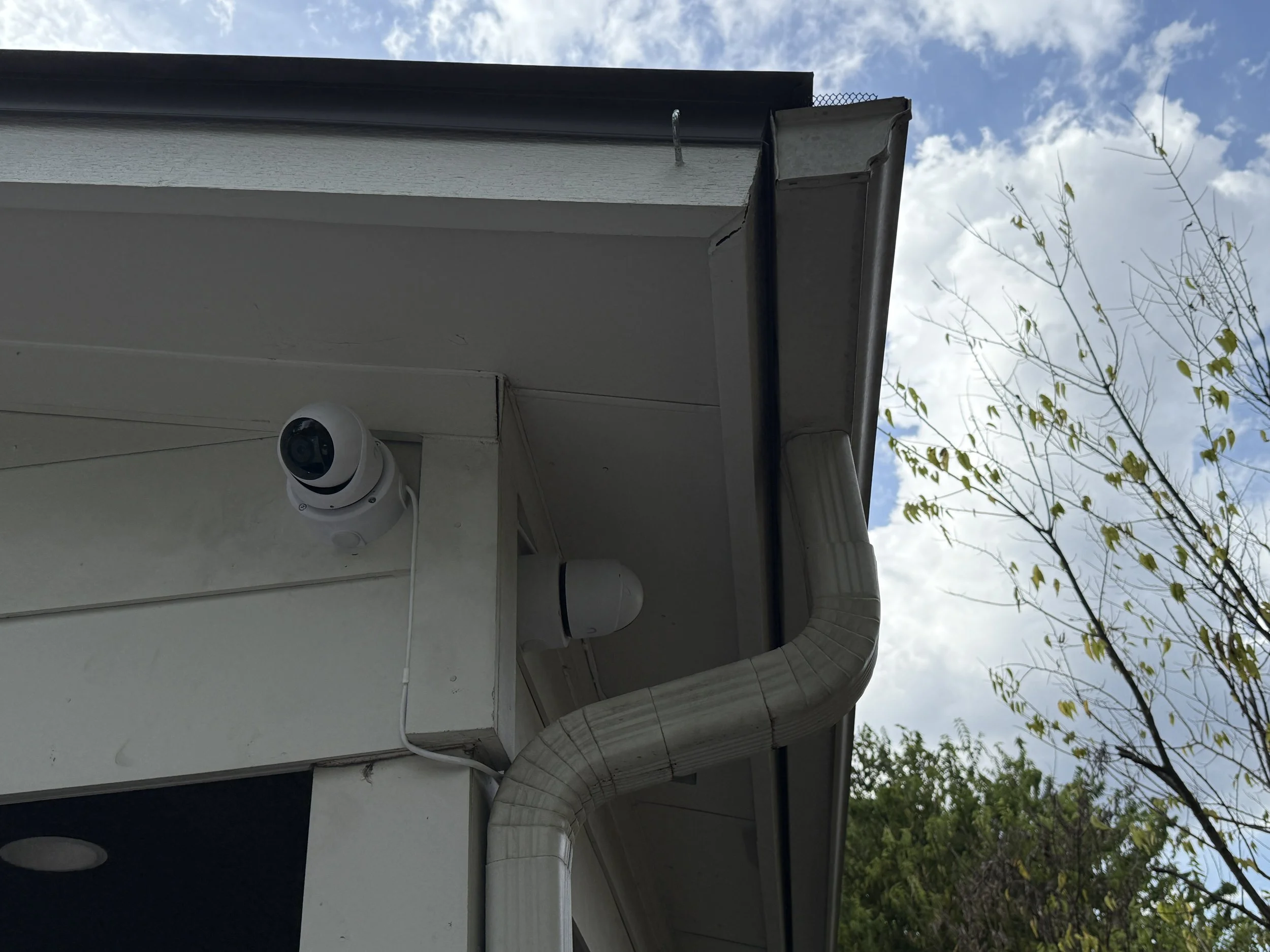 Security camera mounted on the exterior wall of a house near the roof with a rain gutter, against a background of skyline with clouds and trees.
