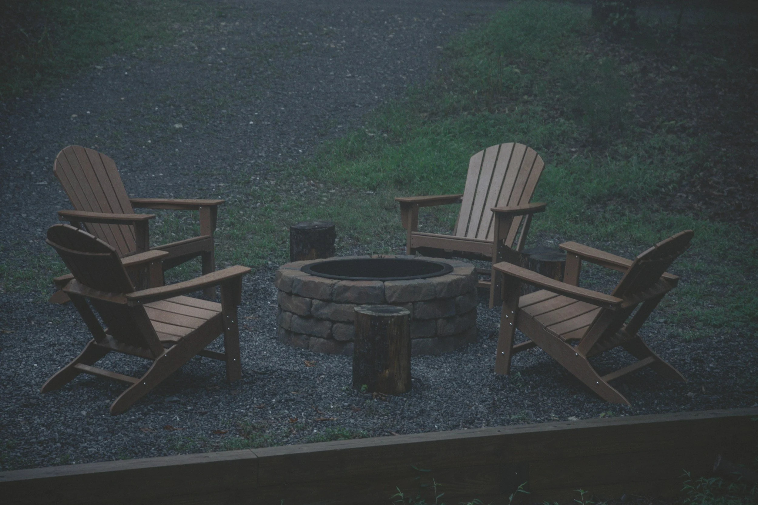 Four Adirondack chairs arranged in a circle around a stone fire pit outdoors, with a grassy and gravel area in the background.