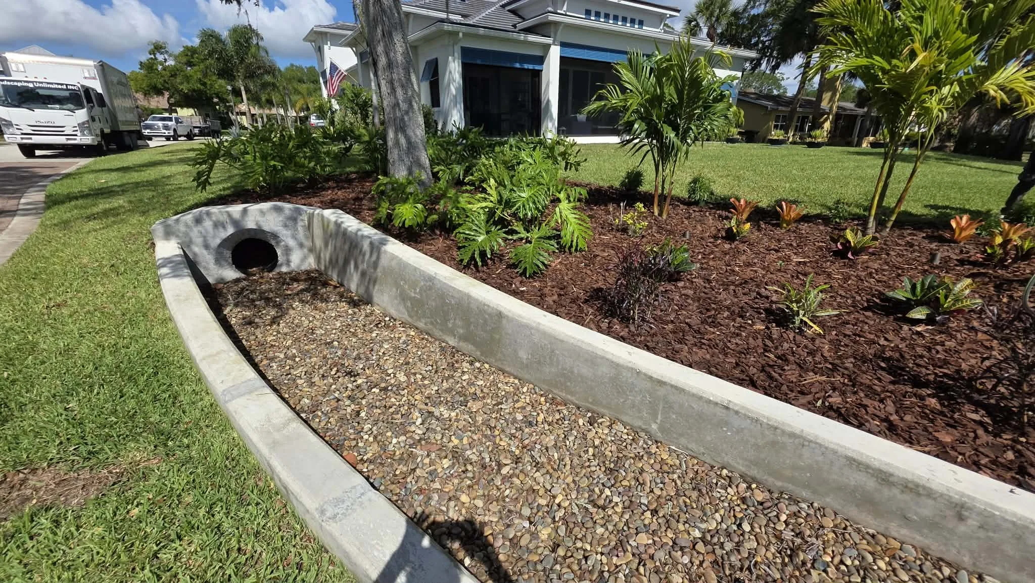 Front yard landscaping with a gravel border, small plants, and mulch, in front of a white house with a porch, several trees, and parked vehicles along the street.