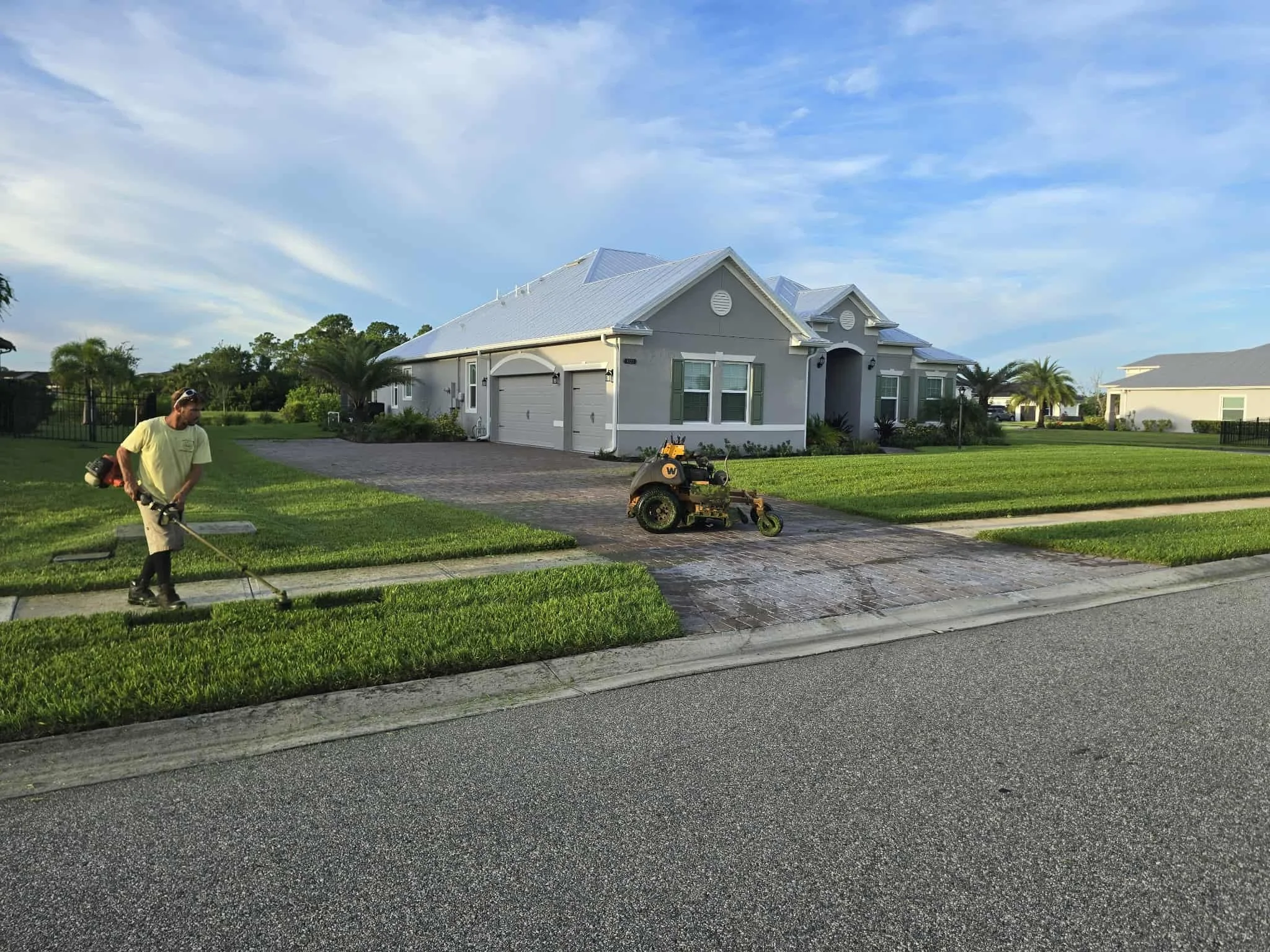 A man working on a lawn mower outside a light-colored house with green shutters, a driveway, and a lawn, under a partly cloudy sky.