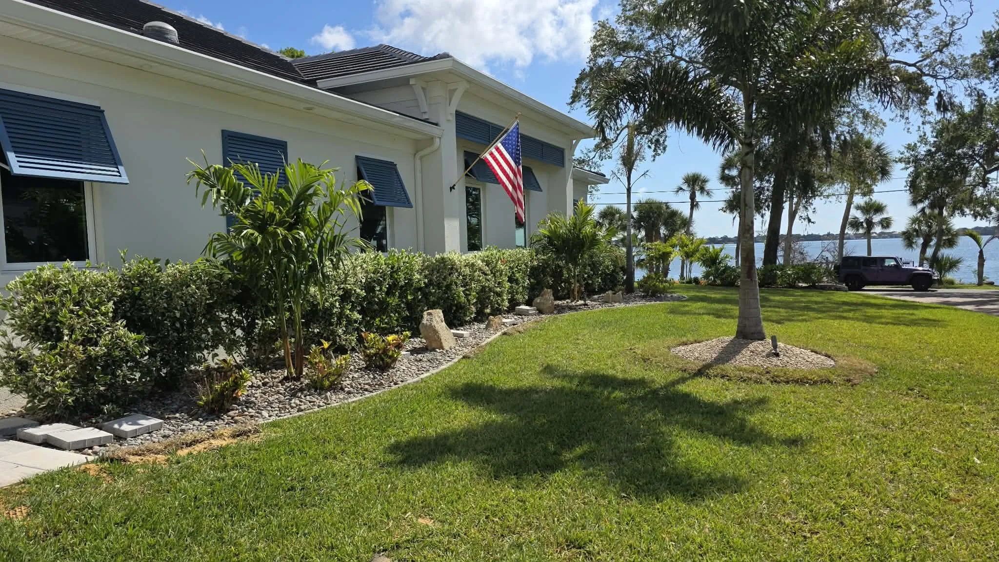 A house with open blue shutters, an American flag, a landscaped yard with grass and bushes, palm trees, and a body of water in the background.