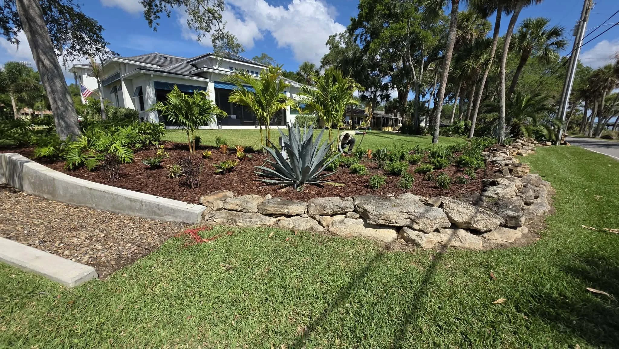A landscaped front yard with a variety of tropical plants, a stone border, and a two-story house with a porch and blue window awnings. The sky is partly cloudy and there are palm trees along the street.