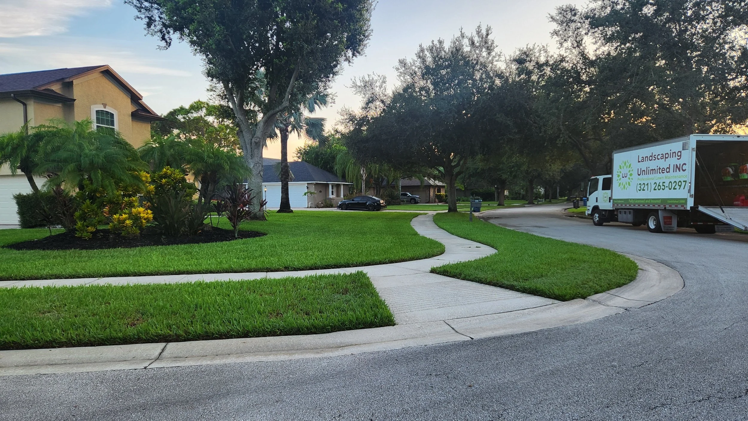 A suburban neighborhood with well-maintained lawns, trees, and houses. A landscaping truck parked on the driveway, and a sidewalk curves through the yards during early evening.