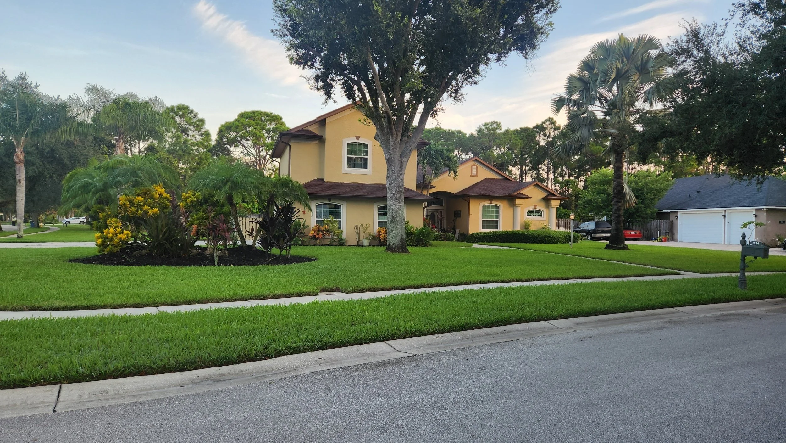A yellow two-story house with a brown roof, surrounded by lush green grass, trees, and bushes. There are several trees including palm trees, and a driveway with parked cars. The sky is partly cloudy with some sunlight.
