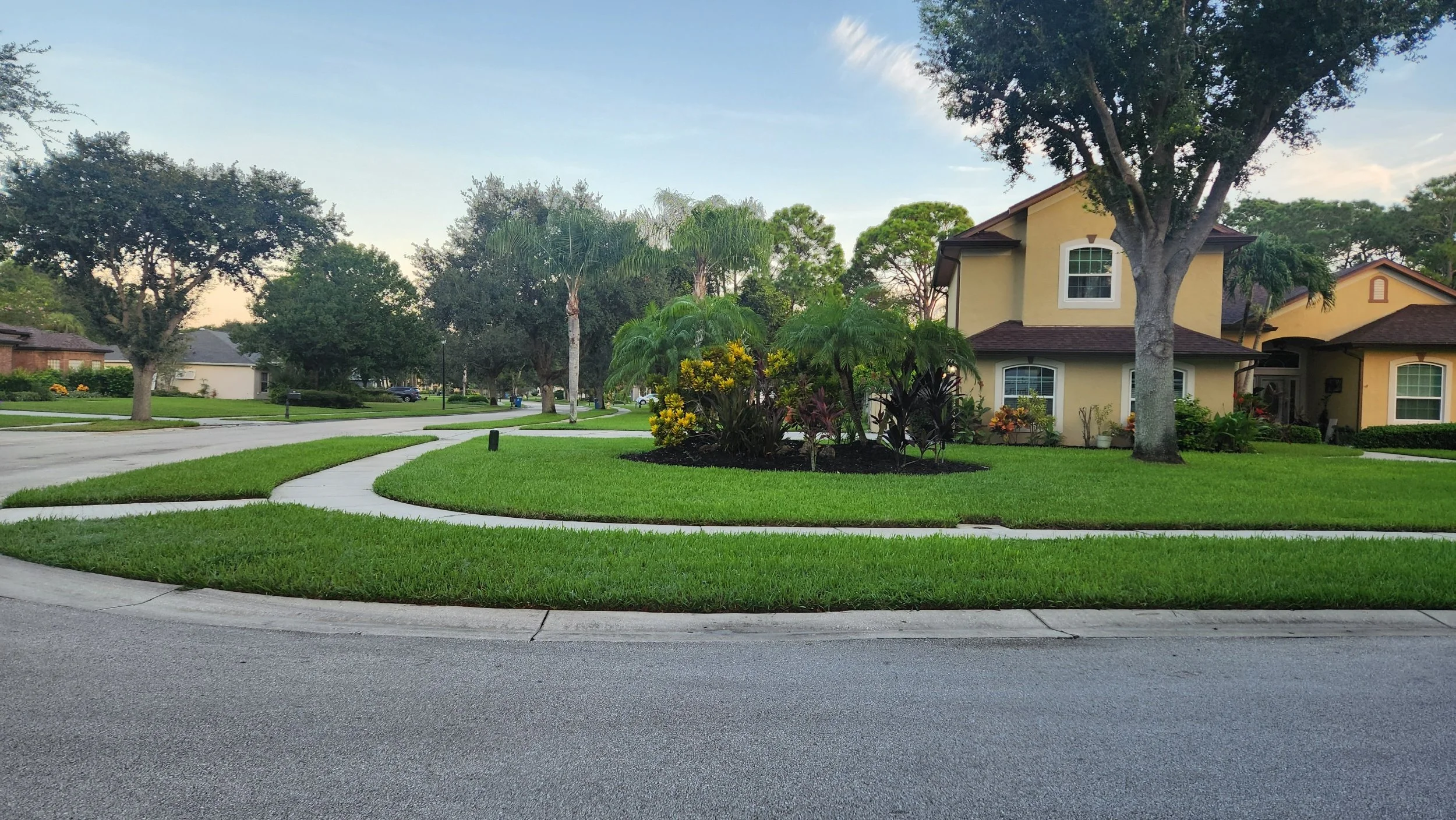 A suburban neighborhood with a yellow house, trees, and manicured lawn, under a partly cloudy sky.